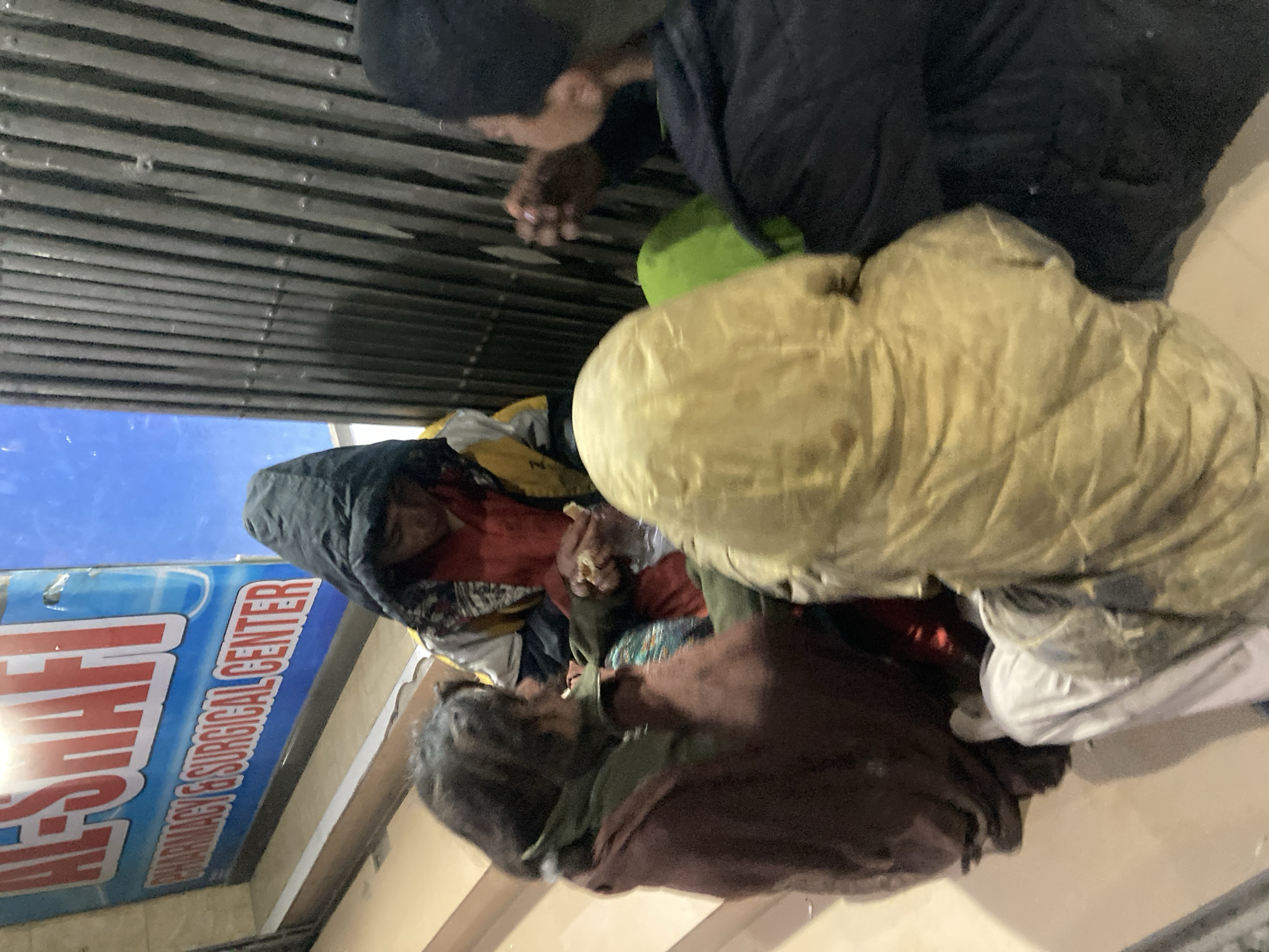 In Quetta City Tehsil, Pakistan, several individuals are huddled on an outdoor staircase adjacent to a pharmacy sign reading "AFSHAFI Pharmacy & Surgical Center." One person is wrapped in a tan-colored sleeping bag or heavy blanket. A second person, wearing a black hooded jacket, is visible partially, their face obscured by the hood. Another individual, covered in a dark brown blanket, sits closest to the bottom of the stairs. Another person, in a black jacket and cap, appears to be interacting with the group. The setting suggests an urban environment, potentially an overnight shelter location.