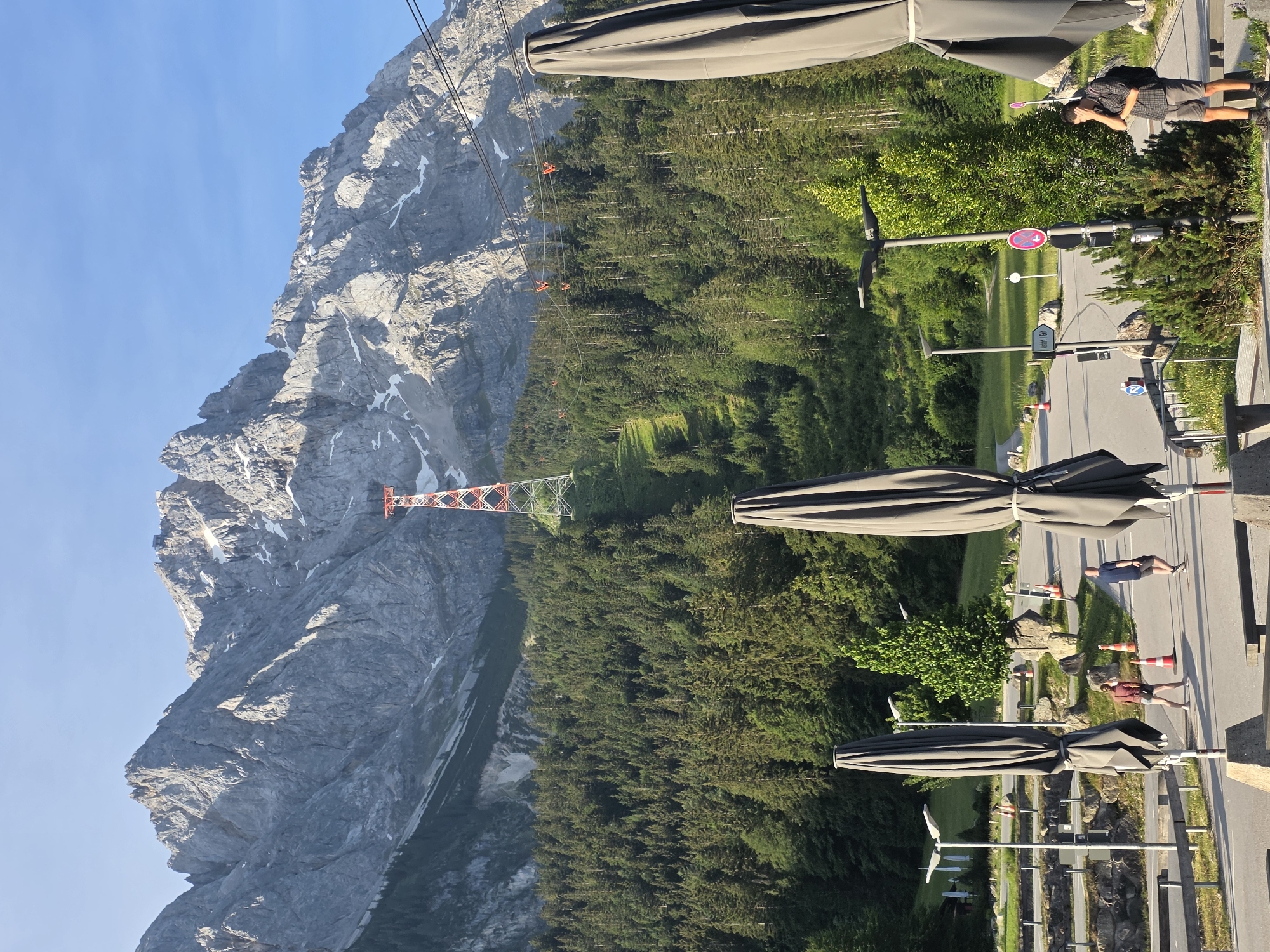 The image depicts a scenic view in Grainau, Germany, dominated by a towering mountain range under a clear blue sky. In the foreground, large, closed grey umbrellas stand tall, casting shadows. A paved area with streetlights and traffic cones is visible, with two individuals walking. One person, a male wearing a dark t-shirt and shorts, walks away from the camera, while another person, a female in athletic wear, walks towards the camera. The middle ground is filled with a dense forest of evergreen trees, stretching up the mountainside. A prominent red and white pylon for a cable car system stands out against the green foliage and rocky mountain slopes. The mountain itself is rugged and imposing, with patches of snow visible near its summit. The overall atmosphere is tranquil and summery, with the sunlight illuminating the landscape.