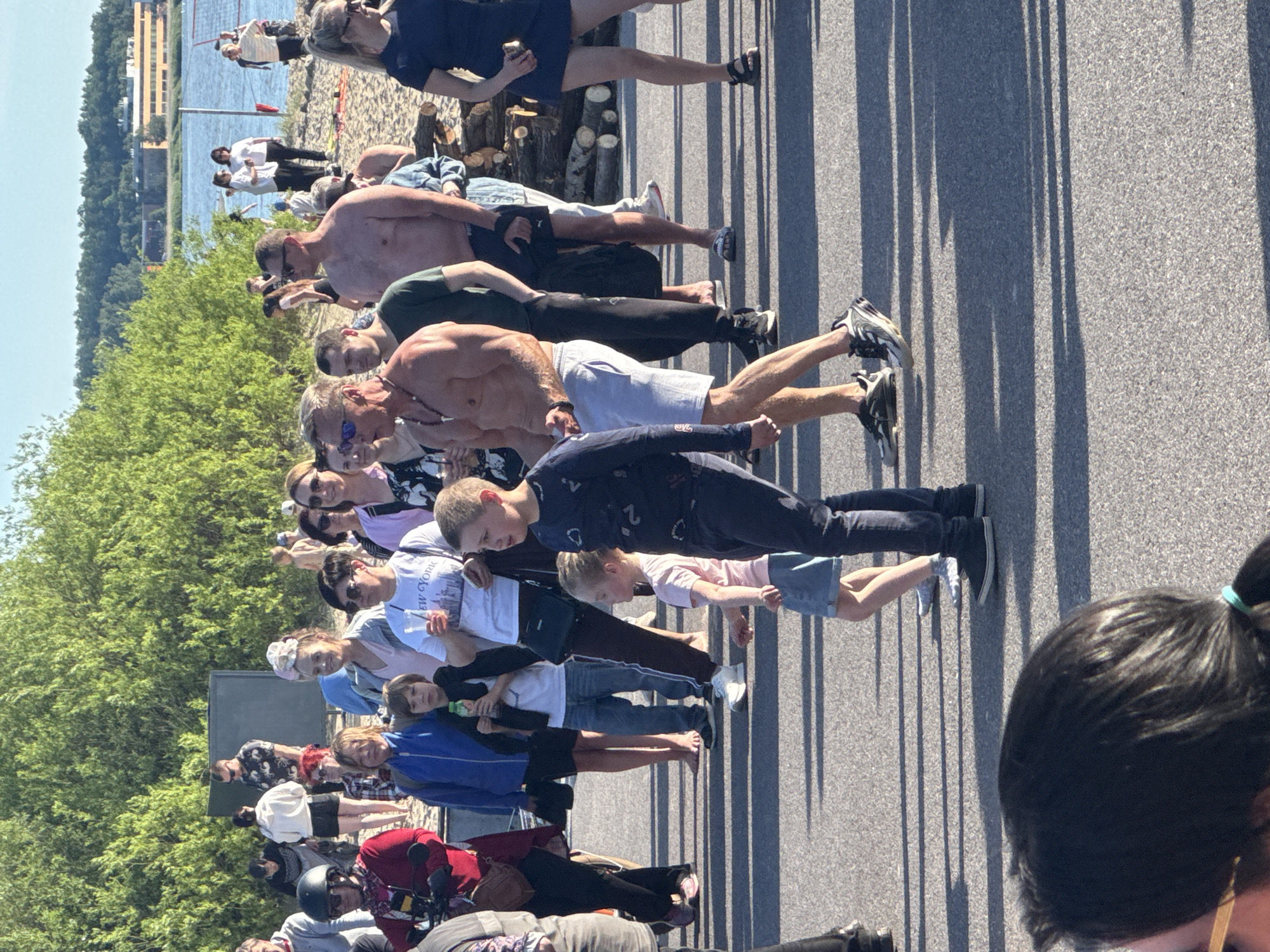 The image shows a group of people gathered on an asphalt surface near a body of water, possibly a river or bay, with a shoreline and trees in the background. The scene appears to be outdoors during the daytime, with sunlight casting long shadows.
In the foreground, a young boy in a dark blue shirt and jeans is walking, looking to his right. To his left and slightly in front of him, a younger girl in a pink shirt and blue skirt is also walking, looking to her left. Behind them, a man with a muscular physique, shirtless and wearing light grey shorts, is looking forward. To his right, a man in a dark green t-shirt and black pants is also looking forward. Further to the right, a person in a dark blue dress is standing and looking to their left.
In the middle ground, a variety of people are present. Some are standing and talking, while others are walking or observing. There is a stack of logs to the right of the center. The background shows more people, trees, and buildings on the far bank of the water, suggesting an urban or semi-urban environment. The presence of a volleyball net in the distance on the sandy shore further indicates a recreational area.
While the exact location in Tallinn, Estonia is not explicitly identifiable from the visual cues, the setting suggests a public gathering place, possibly a waterfront park or beach area, which are common in coastal cities like Tallinn. The overall atmosphere appears relaxed and social.
