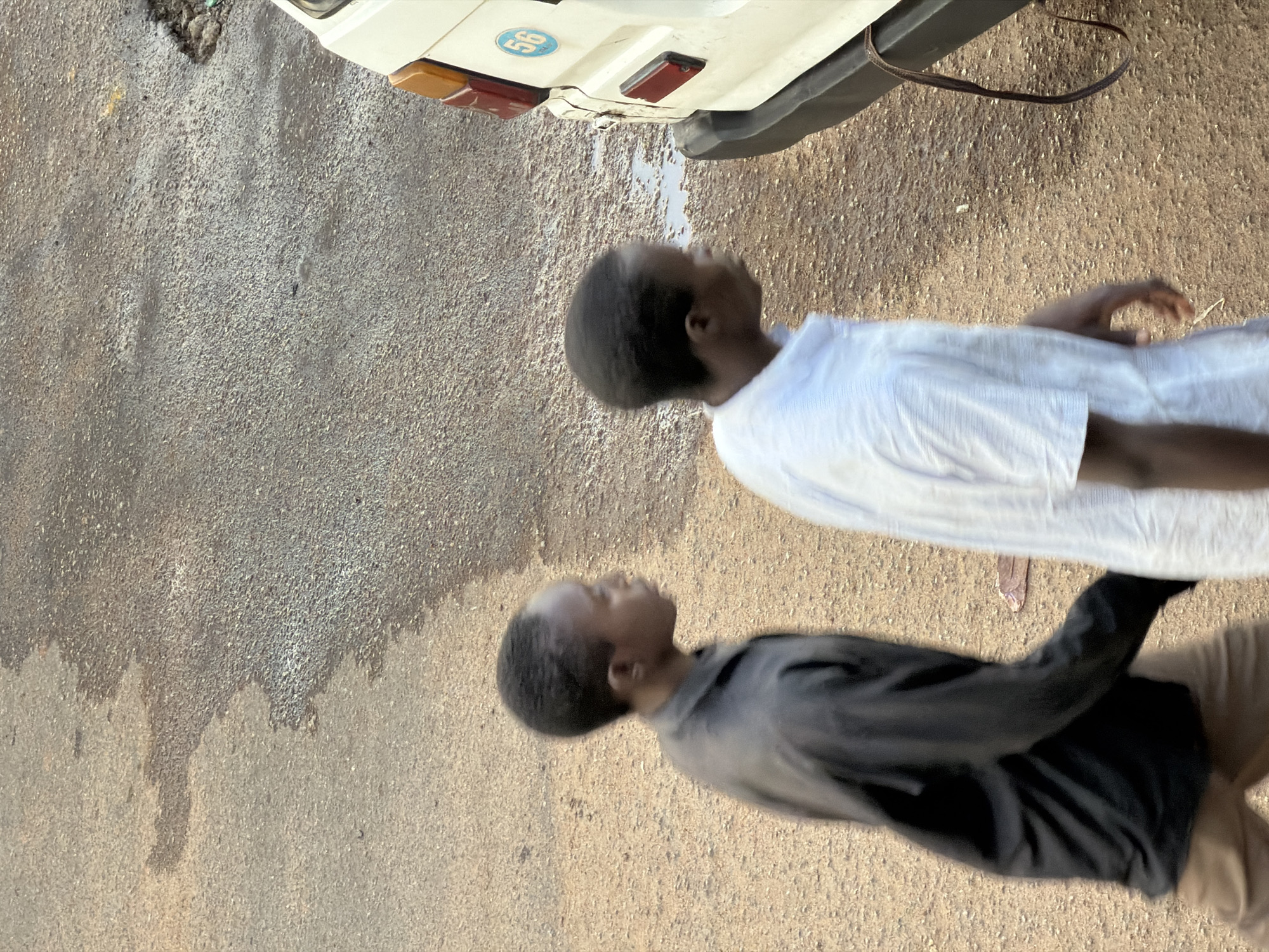 Two young boys, likely of African descent, are standing on a dusty, asphalt road in Bukuru, Nigeria. The boy in the foreground is wearing a dark grey shirt and tan pants. He is looking upwards and to his left, with a neutral expression. The boy behind him is wearing a white shirt and is also looking upwards and to his left. He appears to be slightly taller than the first boy.
The setting is outdoors, with the ground being a mix of grey asphalt and lighter brown soil. There are some dark patches on the ground that could be oil stains or wet spots. In the background, the rear of a white vehicle is visible, with a brown strap hanging from it. The vehicle has a red tail light and a blue circular sticker with the number "56" on it. The sun appears to be high in the sky, suggesting it is daytime. There are no obvious indications of specific weather conditions, though the dusty environment might suggest dry weather. The overall impression is a casual, everyday scene.
