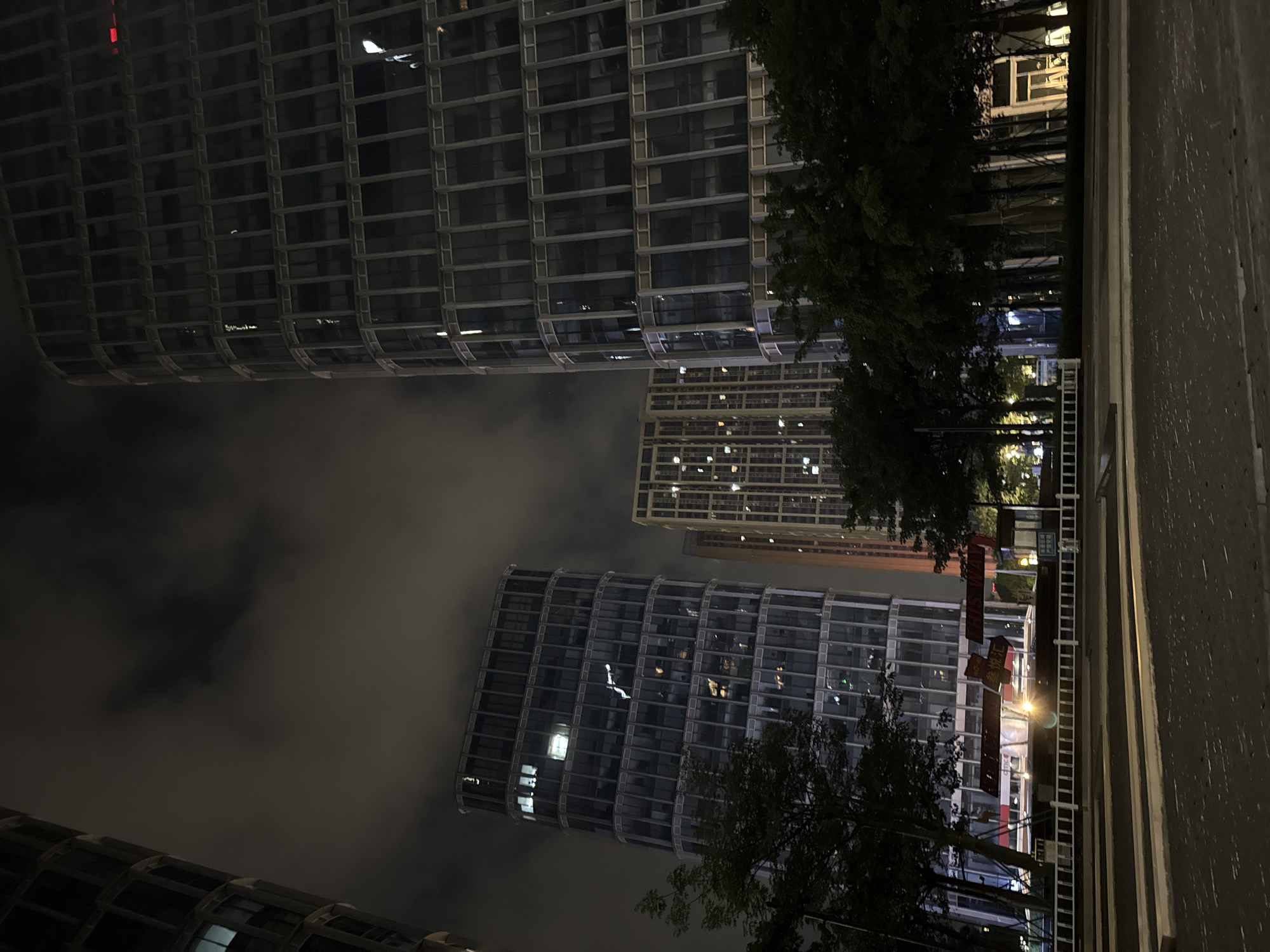 This is a nighttime photograph of a city scene in Haizhu District, China. The image features multiple tall, modern buildings with numerous illuminated windows, suggesting an urban setting with significant human activity. Trees with dark foliage are visible in the foreground and between buildings, adding a natural element to the otherwise architectural landscape. The sky is partially obscured by clouds, which are dimly lit by the city lights, indicating a slightly overcast or hazy night.
The buildings appear to be office or residential complexes, characterized by their grid-like facades and reflective glass exteriors. One building in the foreground has a curved, segmented design. In the background, another building is visible, its windows lit up, suggesting it is also occupied.
A paved area, possibly a sidewalk or street, runs along the right side of the image, bordered by a concrete structure. A white ladder-like structure is attached to the concrete wall, its purpose unclear. Red banners with Chinese characters are hanging between buildings, indicating a local context. The overall mood is one of a bustling, well-lit urban environment at night. There is no clear activity or event taking place beyond the general indication of city life. The time is clearly night due to the darkness and the artificial lighting.