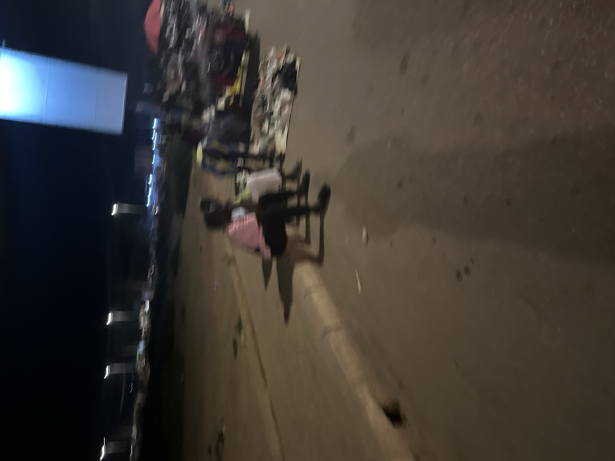 The image depicts a nighttime scene in a market area of Bukuru, Nigeria. Two individuals are seated on a curb, with one wearing a pink top and the other a dark top. To their right, several other people are standing or seated around what appears to be a market stall displaying various items, possibly clothing, on the ground. In the background, there are lights and structures, suggesting an urban or semi-urban environment. A large, illuminated billboard is visible in the upper right portion of the frame. The ground is paved with asphalt, and a drainage culvert is present on the left side of the curb. The overall lighting is dim, typical of a nighttime street scene.
