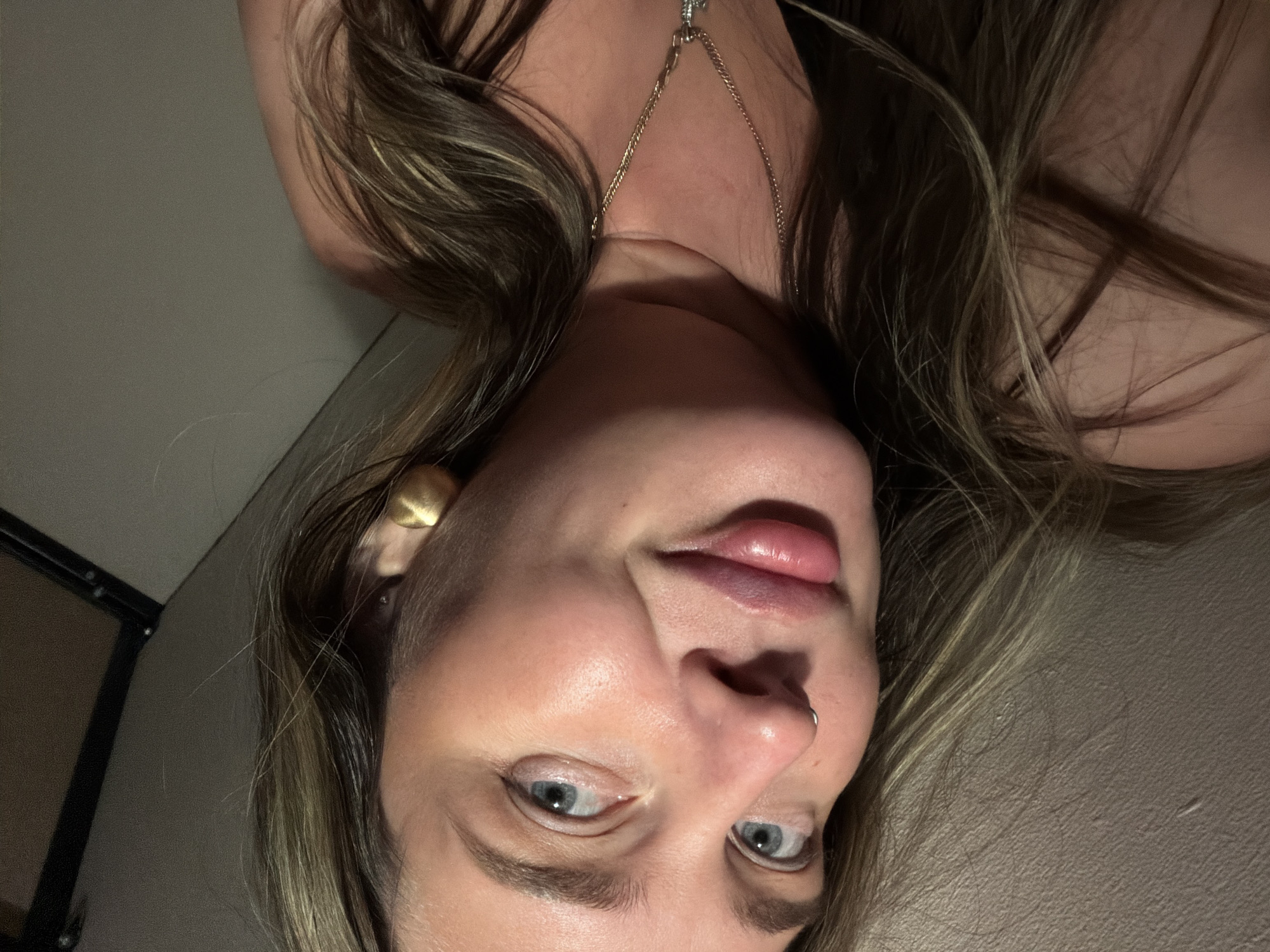 A close-up, upside-down shot features a fair-skinned person with light brown hair. The subject has blue eyes, a nose piercing, and wears a gold earring and necklace. The background is a plain, dark wall with a hint of a dark object on the left side. No location details are available.