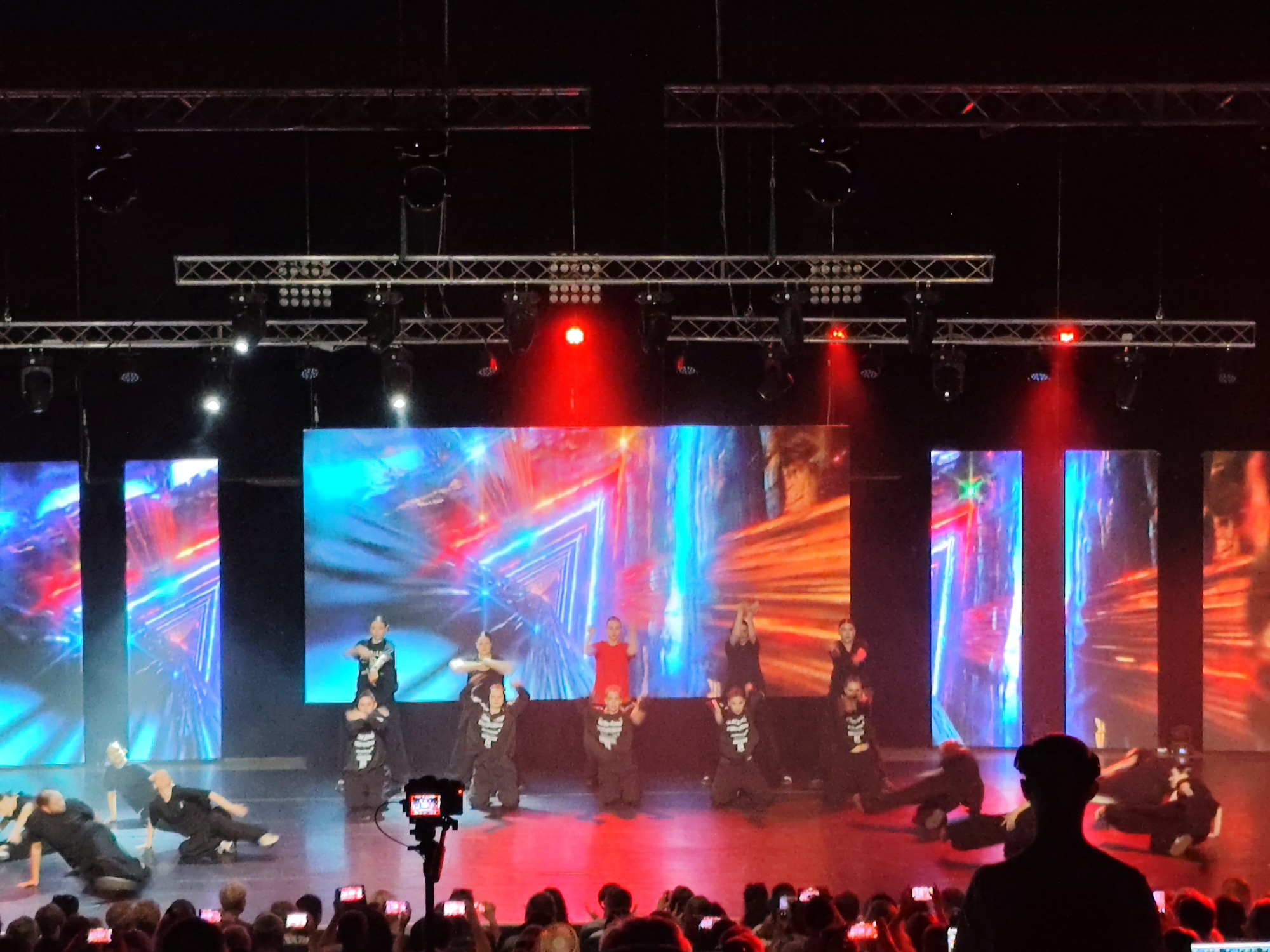The image depicts a dance performance on a stage with a large digital backdrop displaying abstract, colorful, and dynamic graphics. Several dancers, mostly young children and teenagers, are performing various dance moves on the illuminated stage floor. Some dancers are dressed in black outfits, while others wear costumes with white skeleton-like prints. Red and blue stage lights cast dramatic shadows and illuminate the performers. In the foreground, a camera on a tripod is positioned to record the performance, and the silhouettes of audience members holding up mobile phones are visible at the bottom of the frame. The overall scene suggests a lively and energetic event. There is no information in the image to suggest the location is the city of Mykolaiv, Ukraine.
