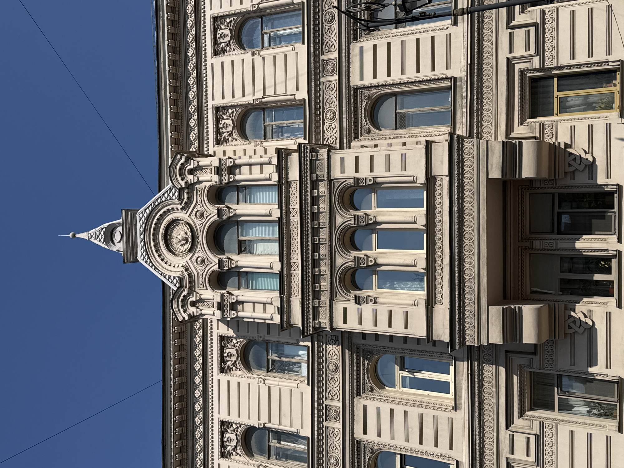 The image shows a close-up, low-angle exterior view of the ornate facade of a historic building in Saint Petersburg, Russia, bathed in sunlight against a clear blue sky. The architecture is highly detailed, featuring intricate decorative moldings, arched windows with multiple panes, and horizontal banding across the building's surface. A prominent central element includes a pedimented section with a circular relief and a pointed spire topped with a finial. Shadows cast by the sun highlight the three-dimensional ornamentation. A single dark electrical wire stretches diagonally across the upper portion of the frame. No people or animals are visible. The building's style suggests it is likely from the late 19th or early 20th century, characteristic of St. Petersburg's architectural heritage.