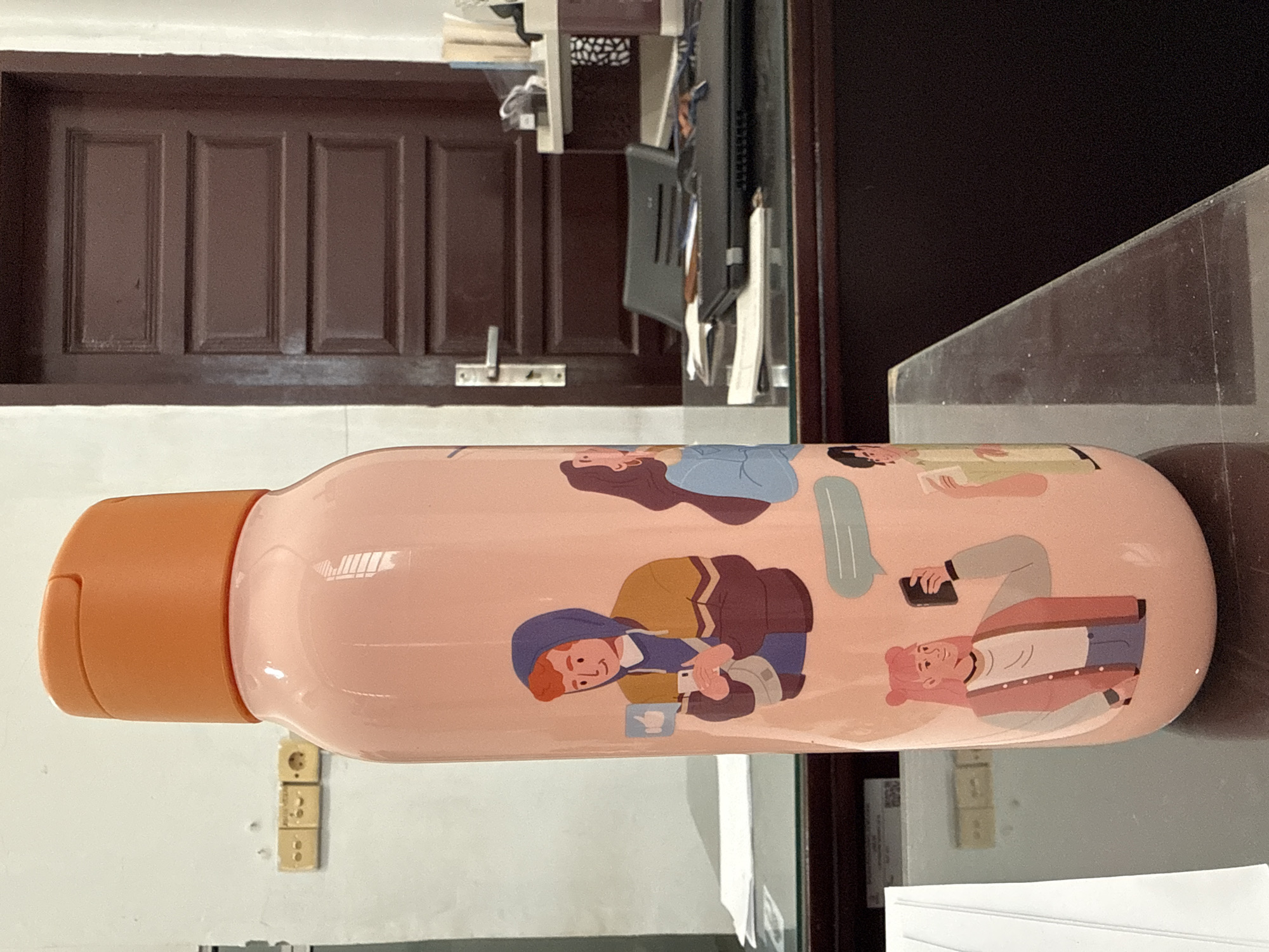 A pink water bottle with orange lid sits on a glossy surface, presumably a desk, in an indoor setting. The bottle is decorated with cartoon illustrations of people. Behind the bottle is a wall with electrical outlets, and a brown wooden door with multiple rectangular panels. In the background, there is a computer monitor and some office supplies, suggesting this might be an office or a home office. Given the electrical outlet style, this photo was taken in Lhokseumawe, Indonesia.