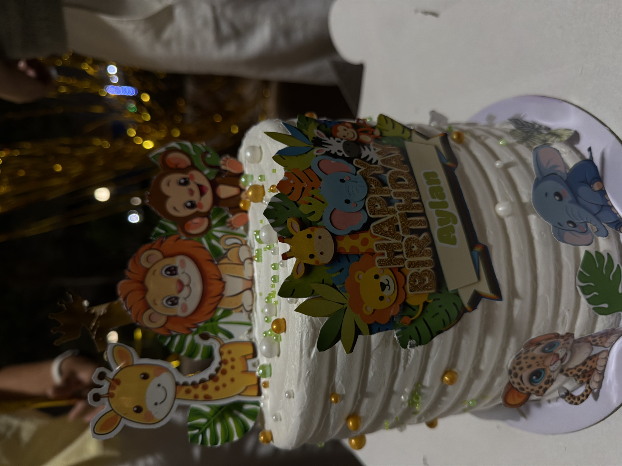 A close-up, eye-level shot captures a white frosted birthday cake adorned with safari-themed decorations. The cake is circular and appears to be made of multiple layers, with a ridged frosting texture. On top of the cake, there are cartoonish figures of a giraffe, a lion, and a monkey, along with green leaves. The front of the cake features a banner that reads "HAPPY BIRTHDAY" in glittery gold letters, with "Aylan" written in green text below it. Surrounding the banner are more cartoon animal figures, including an elephant, a zebra, and another lion. Small edible pearls in white, green, and gold are scattered across the top of the cake.
The cake is positioned on a white cake stand. In the background, out of focus, are individuals with their arms visible, suggesting a celebratory gathering. Golden tinsel curtains hang in the background, creating a festive atmosphere. The lighting is warm and appears to be artificial, with some bokeh effects from the lights. The scene is indoors, likely a home or event space, and does not provide specific clues to its exact location within 6th of October City, Egypt, other than it is a celebration.