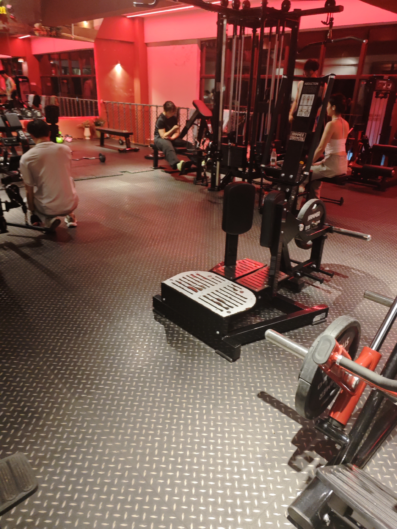 The image shows the interior of a gym, likely located in the Chongqing High-tech Zone, China. The gym has a metallic, diamond-plated floor. The lighting is primarily red. Several people are engaged in various exercises, including one individual using a leg extension machine, another seated on a leg press machine, and a third person near the foreground, kneeling. The gym features a range of equipment, including weight machines, barbells, and a cable crossover system. The overall environment appears to be a contemporary fitness center.