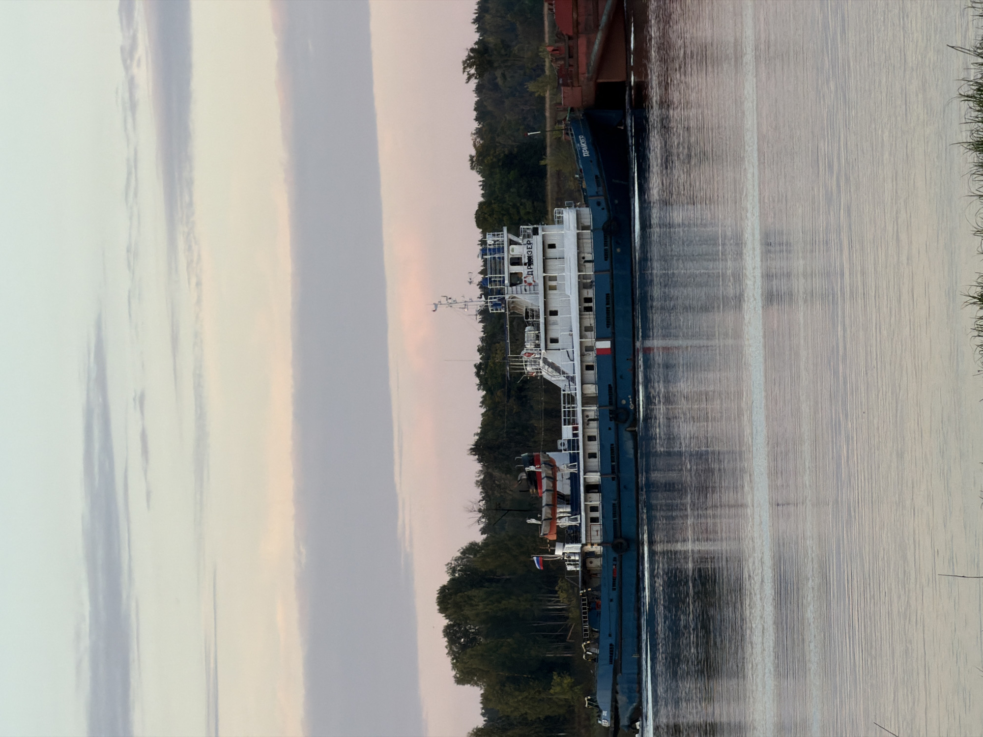 A navy blue and white tugboat travels on a body of water. The tugboat has multiple windows and a red and white life preserver visible on its deck. A Russian flag is also flying on the boat. The boat is pushing a large, red barge. In the background, a dense forest lines the shore under a pale sky with streaks of pink and purple clouds. The water reflects the sky and the boat. The scene appears to be set in Chernushka, Russia.