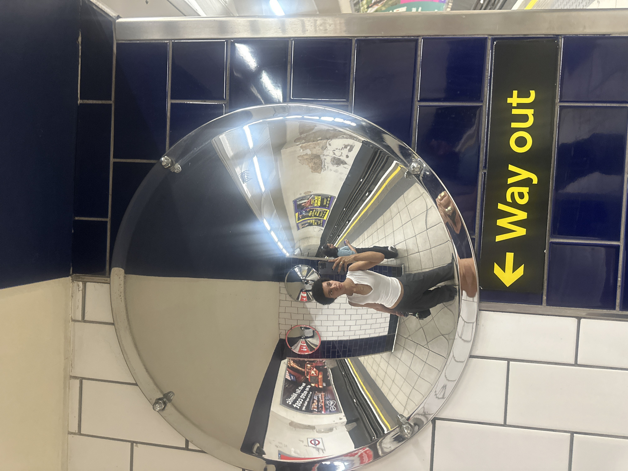 The image shows a person taking a selfie in a London Underground station. The person is standing in front of a large convex mirror, which reflects the station platform. The platform has tiled walls, some of which are white and some are dark blue. There is a sign with a yellow arrow pointing to the left and the words "Way out" in yellow lettering. In the reflection, another person can be seen in the background, as well as advertisements on the wall of the tunnel. The person taking the selfie is wearing a white tank top and dark pants. The overall scene depicts a typical London Underground station environment.