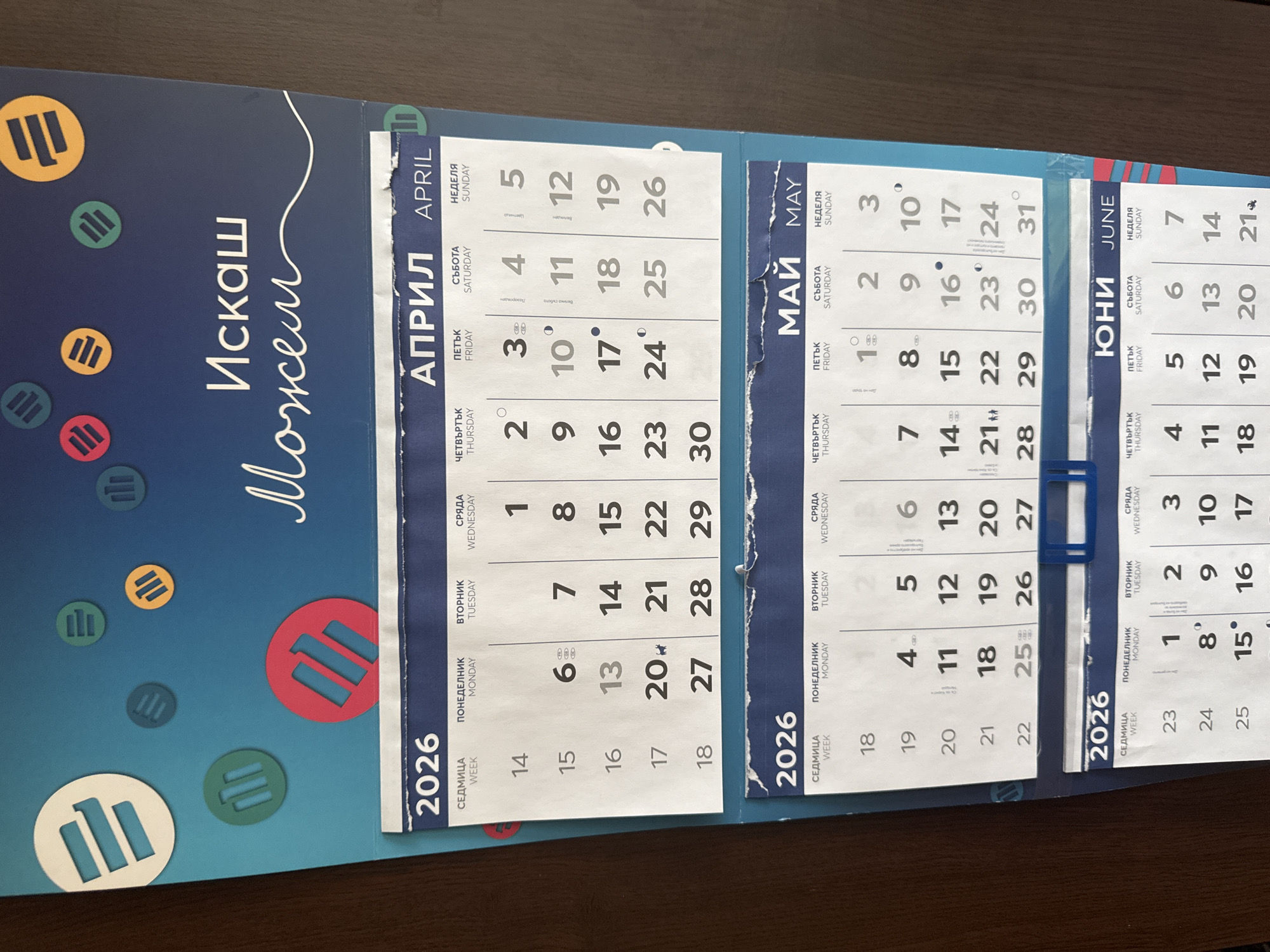 The image shows a section of a wall calendar for the year 2026. The calendar features two months: April and May. Each month is presented in a grid format, displaying the days of the week in Bulgarian and English at the top, followed by the corresponding dates. The calendar has a blue background with stylized circular icons scattered across the top portion. The overall scene is a close-up shot of the calendar, indicating it is likely hanging on a wall indoors. There are no people visible in the image. The calendar's design appears modern and organized. The image does not provide any information that would allow for the identification of a specific location within the city of Sofia, Bulgaria.