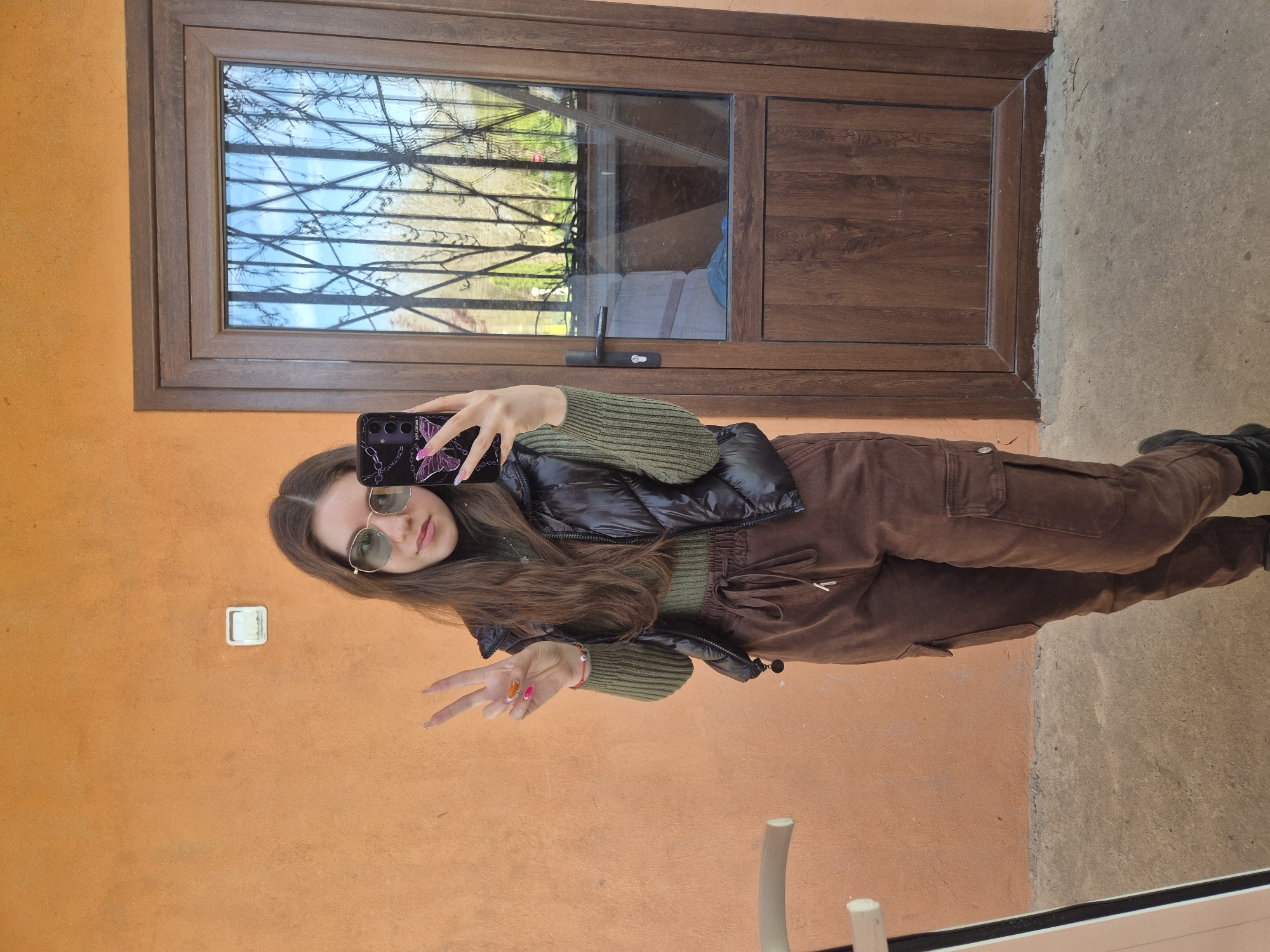 A young woman is taking a selfie in what appears to be an outdoor setting. She has long brown hair and is wearing sunglasses, a green sweater, a black puffer vest, and brown cargo pants. She is holding a phone with a butterfly-themed case and making a peace sign with her hand.
The background consists of an orange wall with a light switch. There's a large brown-framed window/door with visible foliage outside and some reflected furniture inside. The ground appears to be concrete or pavement.
While not explicitly stated, the query requests a location in Pavlikeni, Bulgaria. Assuming the image was taken there, the specific location cannot be determined from the image alone, but the scene appears to be a residential or commercial property.