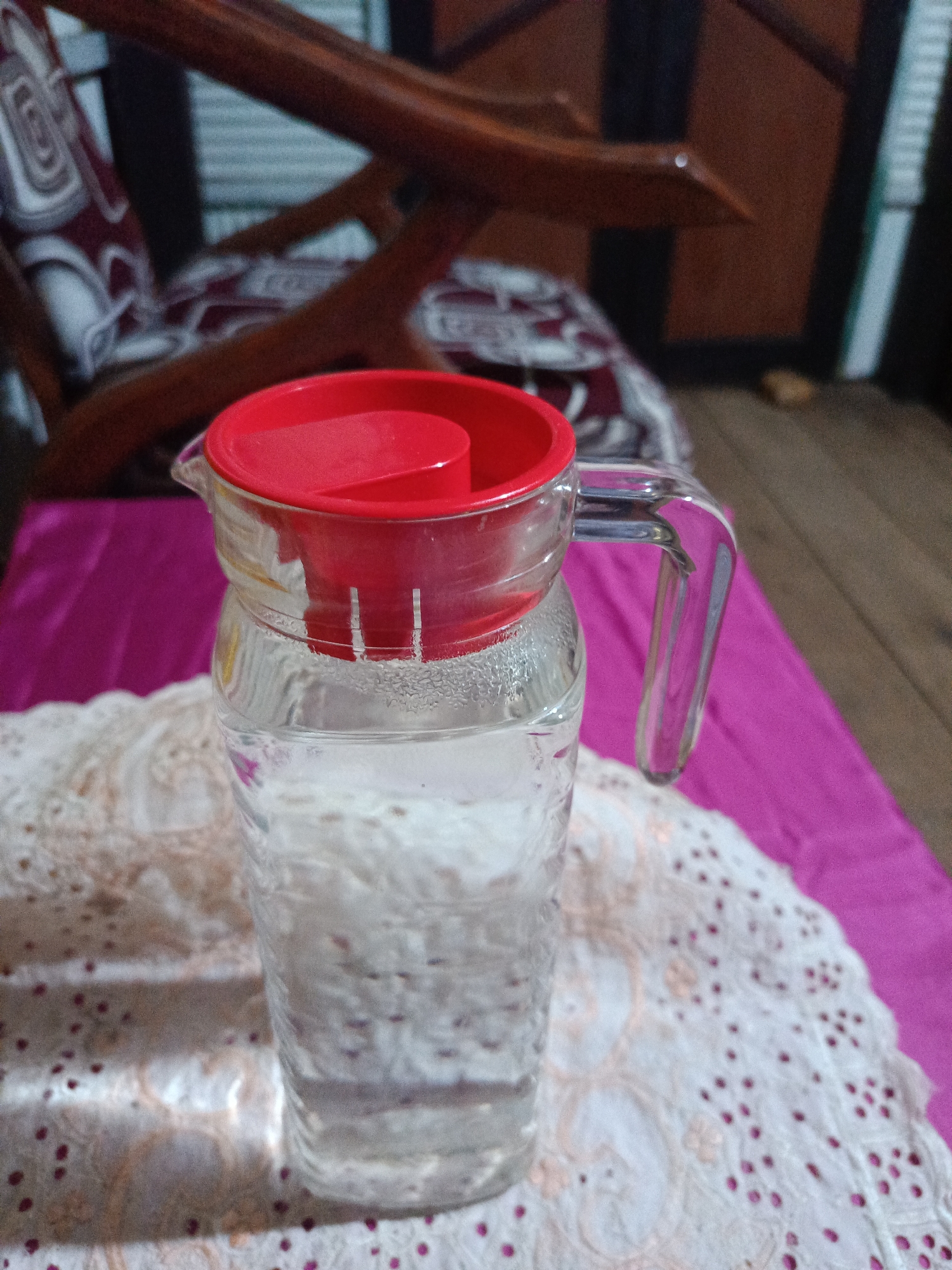 A glass pitcher filled with clear liquid, topped with a red lid, rests on a white lace doily on a pink tablecloth. The pitcher has a clear glass handle and a spout. In the background, a wooden chair with a patterned cushion and a dark wooden door are visible. The scene appears to be indoors, possibly in a home environment within the city of Arateng, Indonesia.