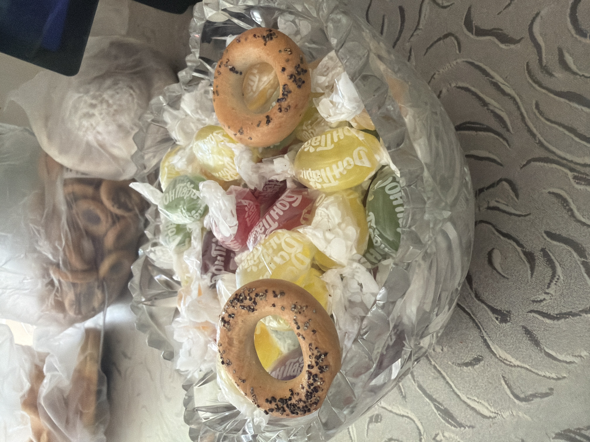 This overhead shot showcases a clear, faceted glass bowl filled with an assortment of candies and baked goods. The candies, individually wrapped in crinkled cellophane, display a variety of bright colors, including yellow, green, and red. Some wrappers reveal text, with "Don Tapi" and "OH!!TA" partially visible. Nestled among the candies are two small, ring-shaped baked goods, likely bagels or similar pastries, dusted with dark poppy seeds. To the left of the bowl, more of these baked goods are visible, also enclosed in plastic packaging. The background features a textured, grey surface with a pattern resembling stylized waves or animal stripes. The lighting suggests an indoor setting, possibly during daylight hours. The overall scene presents a collection of treats, perhaps prepared for a snack or a special occasion. The location context provided is Чёрная, Russia, though there are no overt visual cues within the image to confirm this specific location.