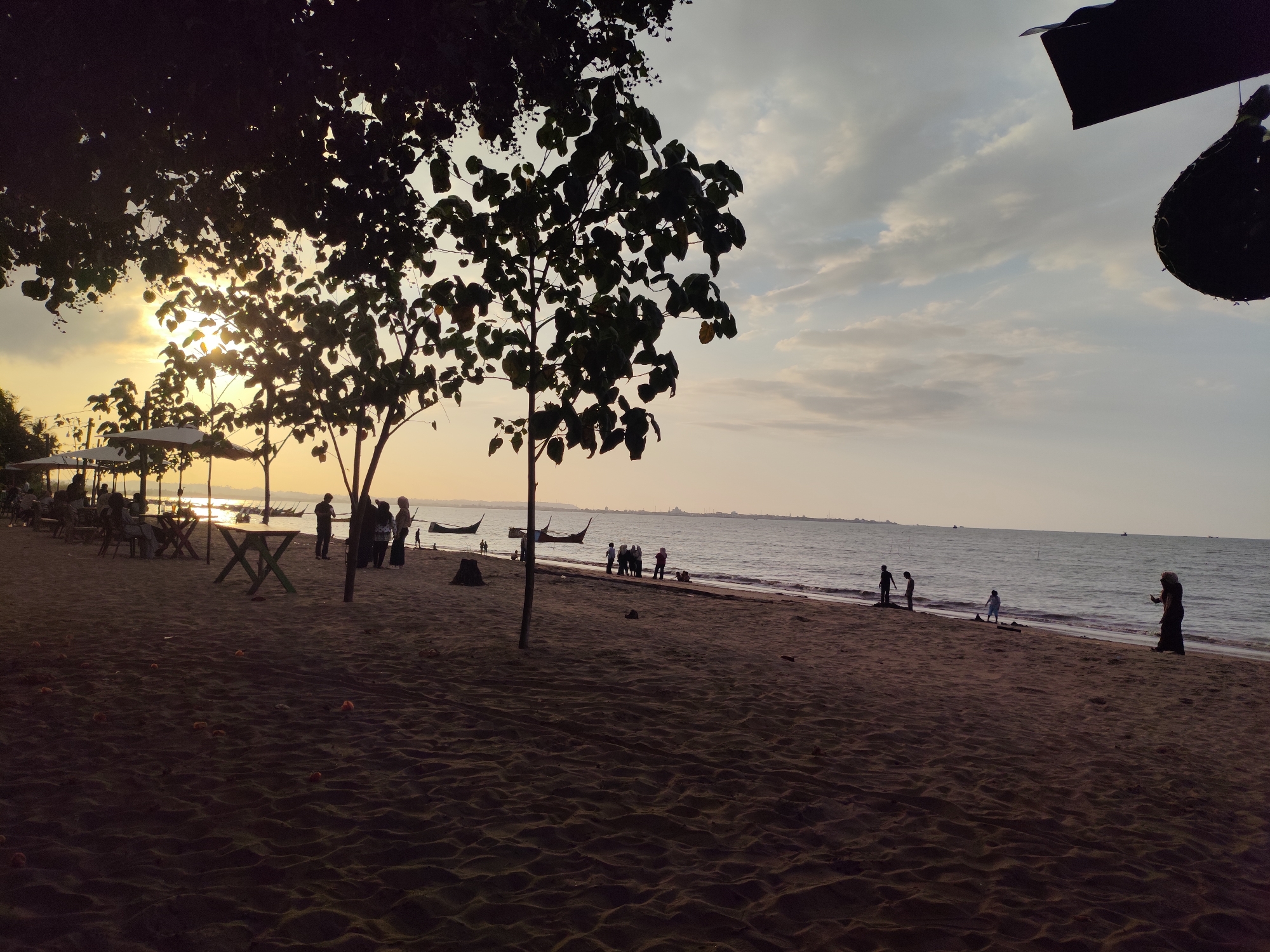 The image depicts a beach scene at sunset in Lhokseumawe, Indonesia. The foreground is dominated by dark, rippled sand, with scattered debris. In the middle ground, several people are visible, some standing on the shore and others walking along the waterline. A few boats are anchored in the sea. The background shows a calm ocean under a sky with soft clouds. Silhouetted trees with lush foliage are prominent in the left side of the image, with some umbrellas and tables suggesting a seating area for beachgoers. The sun is setting, casting a warm, golden light across the sky and water. The overall atmosphere is tranquil and serene.