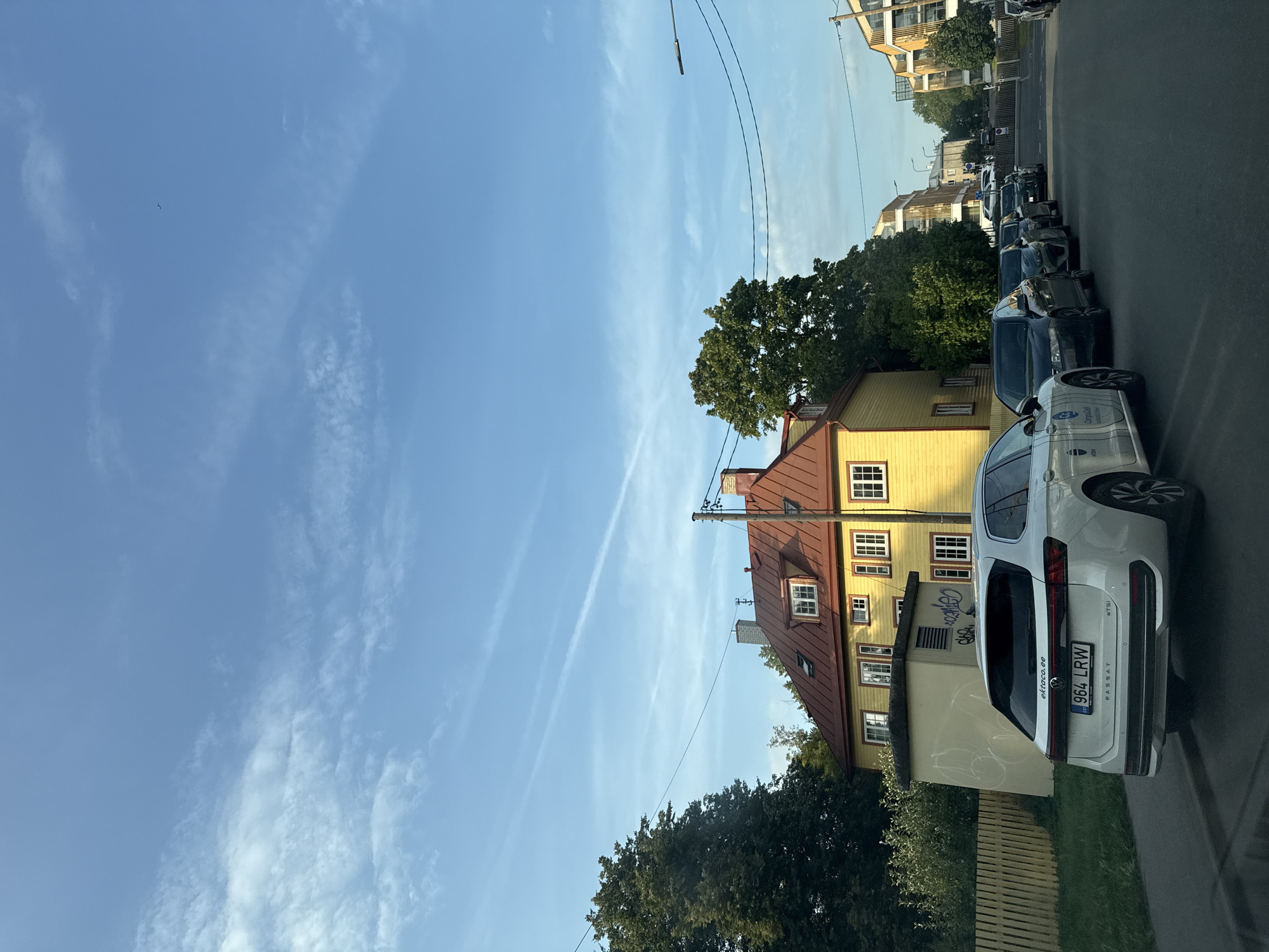 A bright, sunny day in Tallinn, Estonia is depicted in this image. The sky is a clear blue with wispy white clouds. In the foreground, a white Volkswagen Passat station wagon is parked on the right side of the street. Its license plate reads "964 LRW". Behind the Passat, several other cars are parked in a row, including a black sedan. To the left of the street, a yellow house with a red roof stands out, with a wooden fence and lush green trees surrounding it. Further down the street, modern apartment buildings can be seen, suggesting a residential area. The overall scene is one of a quiet, pleasant day in a European city.