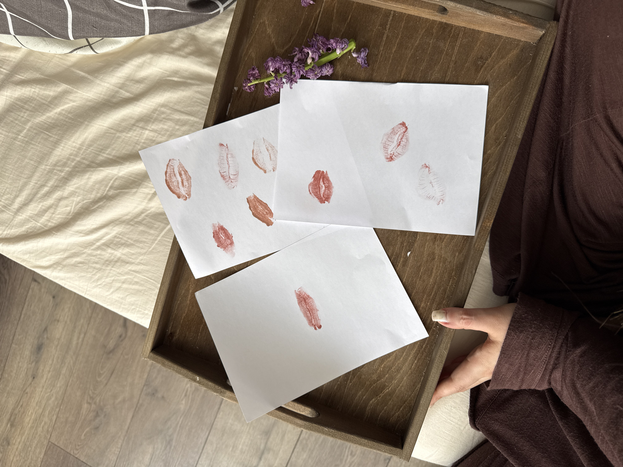 A top-down view depicts a brown wooden tray resting on a bed with neutral-toned linens and a gray and white patterned throw. The tray contains three white pieces of paper bearing lipstick kisses in various shades of red and brown. A small sprig of purple flowers lies beside the papers. The scene appears to be indoors. A person wearing a brown outfit can be seen on the right side of the tray. The scene is located in Krasnodar, Russia.
