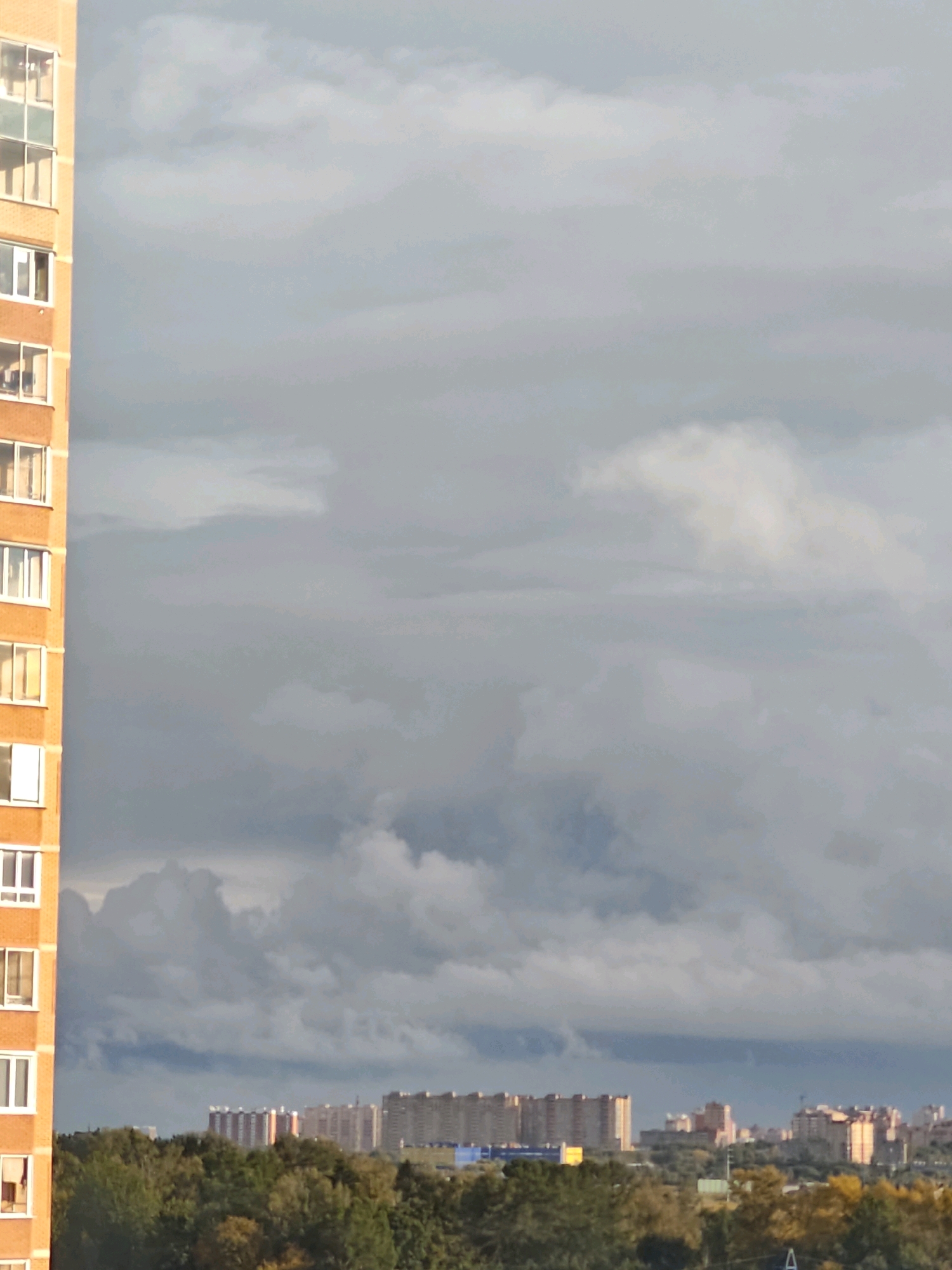 The image shows a view of apartment buildings and trees with a cloudy sky above. On the left, a portion of an orange-colored apartment building with many windows is visible. In the middle distance, a line of multi-story residential buildings stretches across the horizon, some with light-colored facades and others with darker brickwork. A few yellow and orange construction cranes are visible above some of these buildings. The foreground consists of a dense line of green and yellow trees. The sky is filled with a mix of dark gray and lighter white clouds, suggesting overcast weather. The scene appears to be an exterior shot of an urban or suburban landscape.