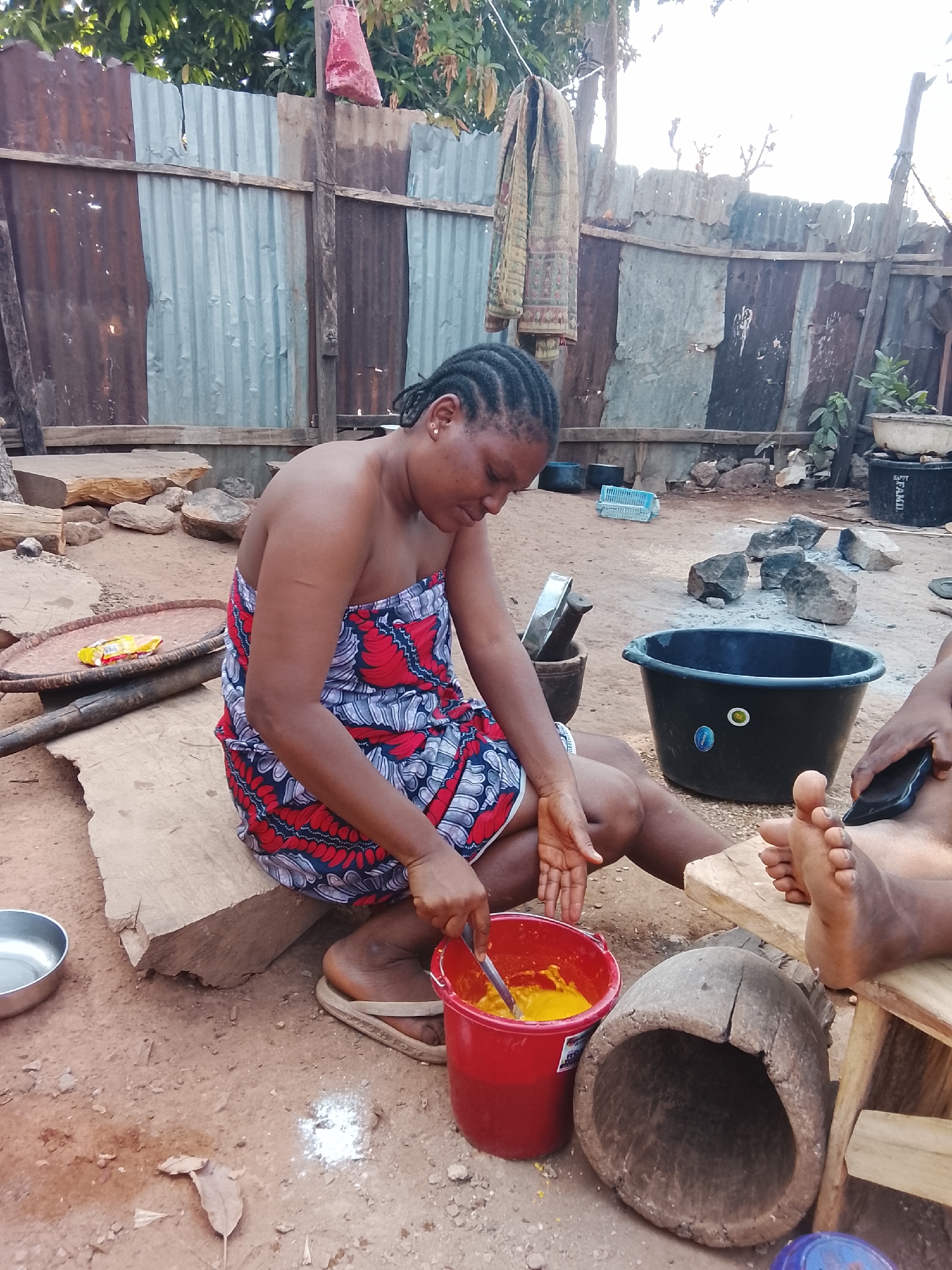 The image shows a woman sitting outside, stirring a yellow substance in a red bucket with a spoon. She is wearing a dress with a red and blue pattern, and sandals. Her hair is braided. She is seated on a large wooden block. A wooden mortar is next to her. A black bucket is in the background, along with rocks and a metal fence. There is another person in the scene sitting next to the woman on a wooden stool. The dirt ground includes a white powder substance and a leaf. The location appears to be a yard with various household items scattered around. The city and region of Unknown, Unknown are noted.