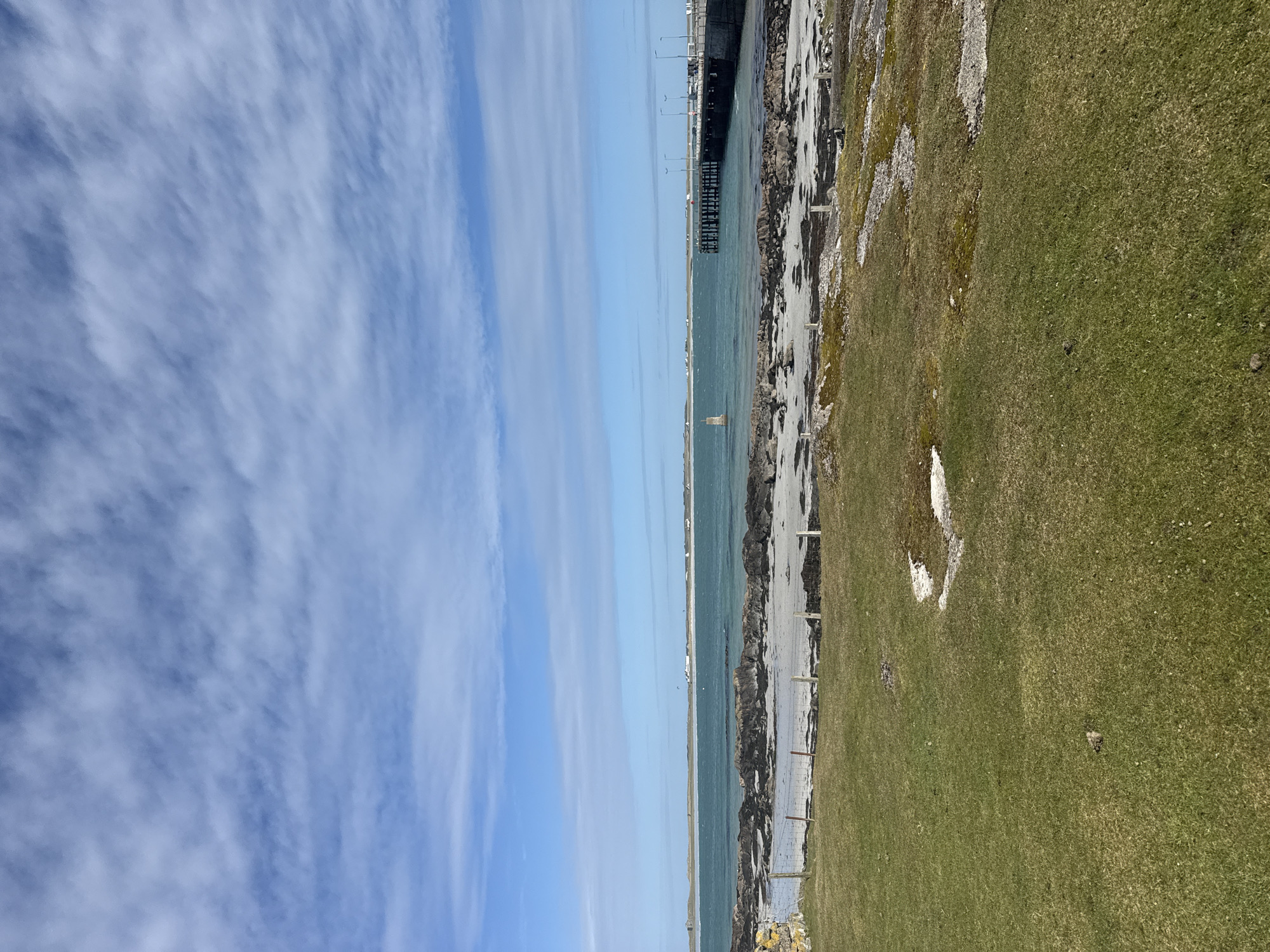 The image depicts a landscape divided into three primary vertical sections: sky, sea, and land.
The sky, on the left, is predominantly blue with wispy, elongated clouds. The sea occupies the central portion, appearing calm with a slight greenish tint. A pier or bridge structure extends into the water.
The land, on the right, is covered in grass, transitioning from green to a mix of brown and sandy areas nearer the water. A fence runs along the land. No people or other identifiable subjects are visible.
The location is Unknown, Unknown.