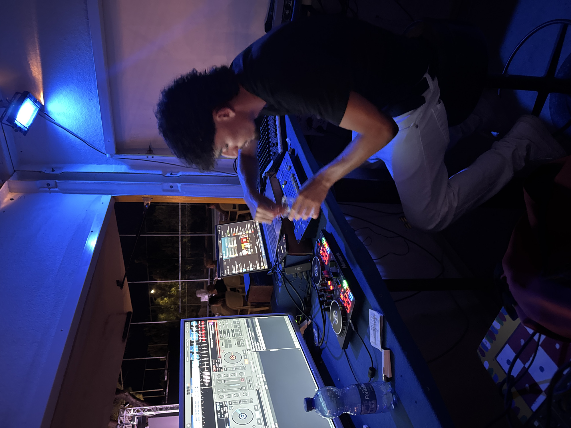 The image depicts a male DJ, with dark, curly hair, operating a DJ setup within what appears to be an outdoor, covered entertainment venue at night. He is wearing a black t-shirt and white pants. In front of him, on a blue table, are two computer monitors displaying DJ software, a laptop, a Numark DJ controller with illuminated buttons, and a bottle of Fontalbe water. The scene is illuminated by blue lighting from above, and a pinkish hue is visible on the right side, suggesting other lighting sources. In the background, partially visible through a fence, are trees and what appear to be other people seated at tables. The setting suggests a casual outdoor event, likely a party or club night. This scene is located in Isola delle Femmine, Italy.