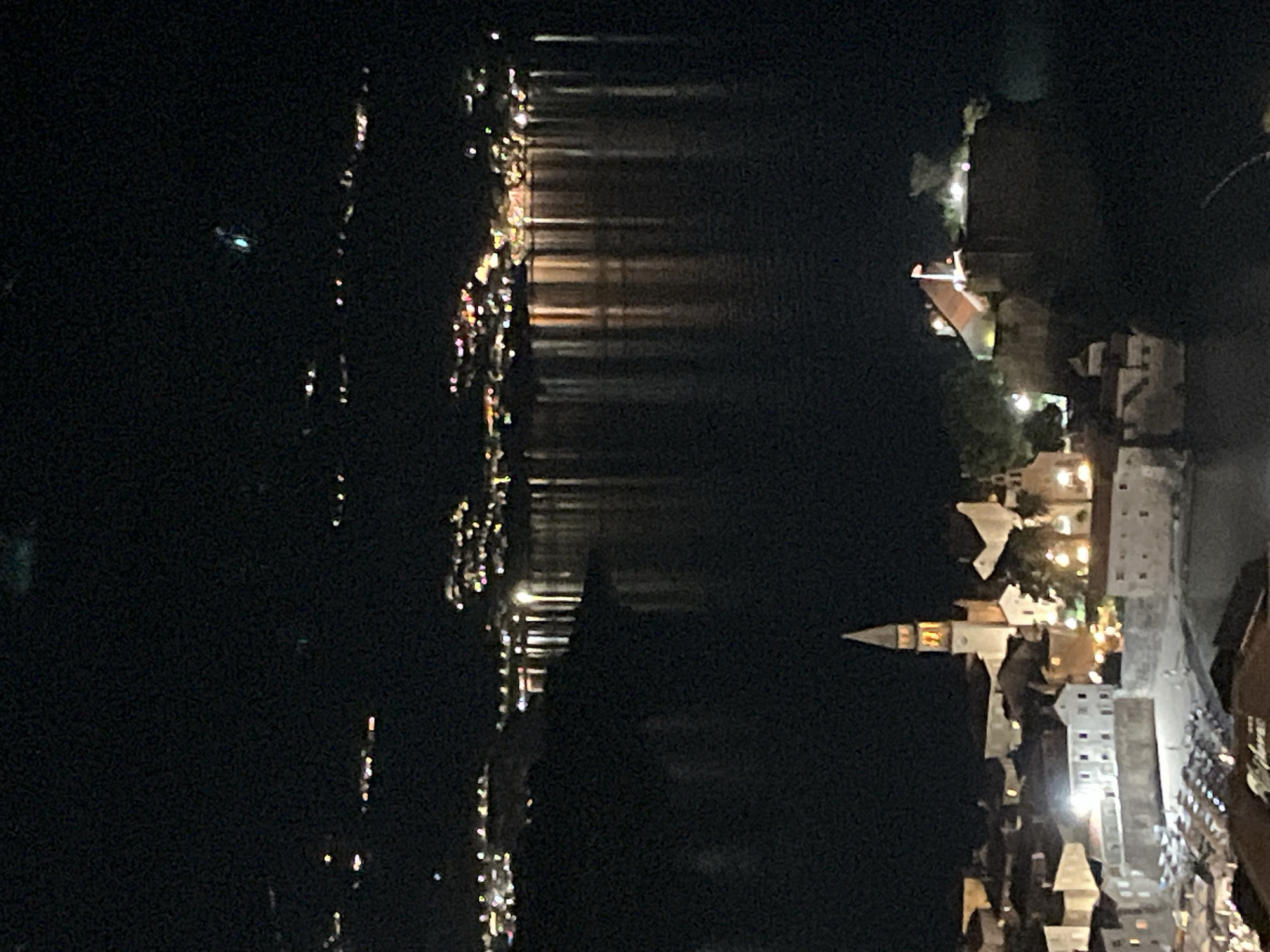 This is a nighttime aerial photograph of the coastal city of Budva, Montenegro. The scene is dominated by the dark expanse of the Adriatic Sea and the illuminated Old Town. In the foreground, the Old Town is visible with its characteristic stone buildings and a prominent church spire. The church is illuminated, with light also emanating from windows of other buildings. A fortified area with a flag on top is also visible on a raised section of the town. In the mid-ground, the calm sea reflects the lights of a distant, illuminated shoreline with buildings clustered on hillsides. The far background shows a hazy, illuminated horizon. The sky above is dark with faint lights visible in the distance.