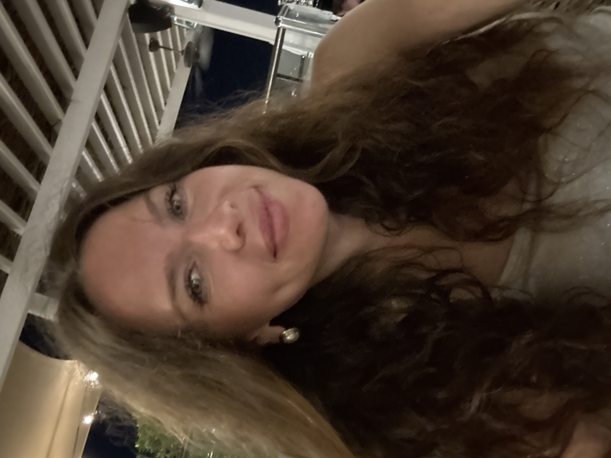 An outdoor selfie taken at night shows a woman with long, wavy brown hair and light eyes. She is wearing a sparkly silver top and a pearl earring in her left ear. She is smiling and looking at the camera. In the background, there is a white slatted ceiling with lights, and further back, a dark sky with what appears to be a ceiling fan and railing. The overall impression is that she is at a restaurant or bar. The specific location in Dubai is not discernible from the image.