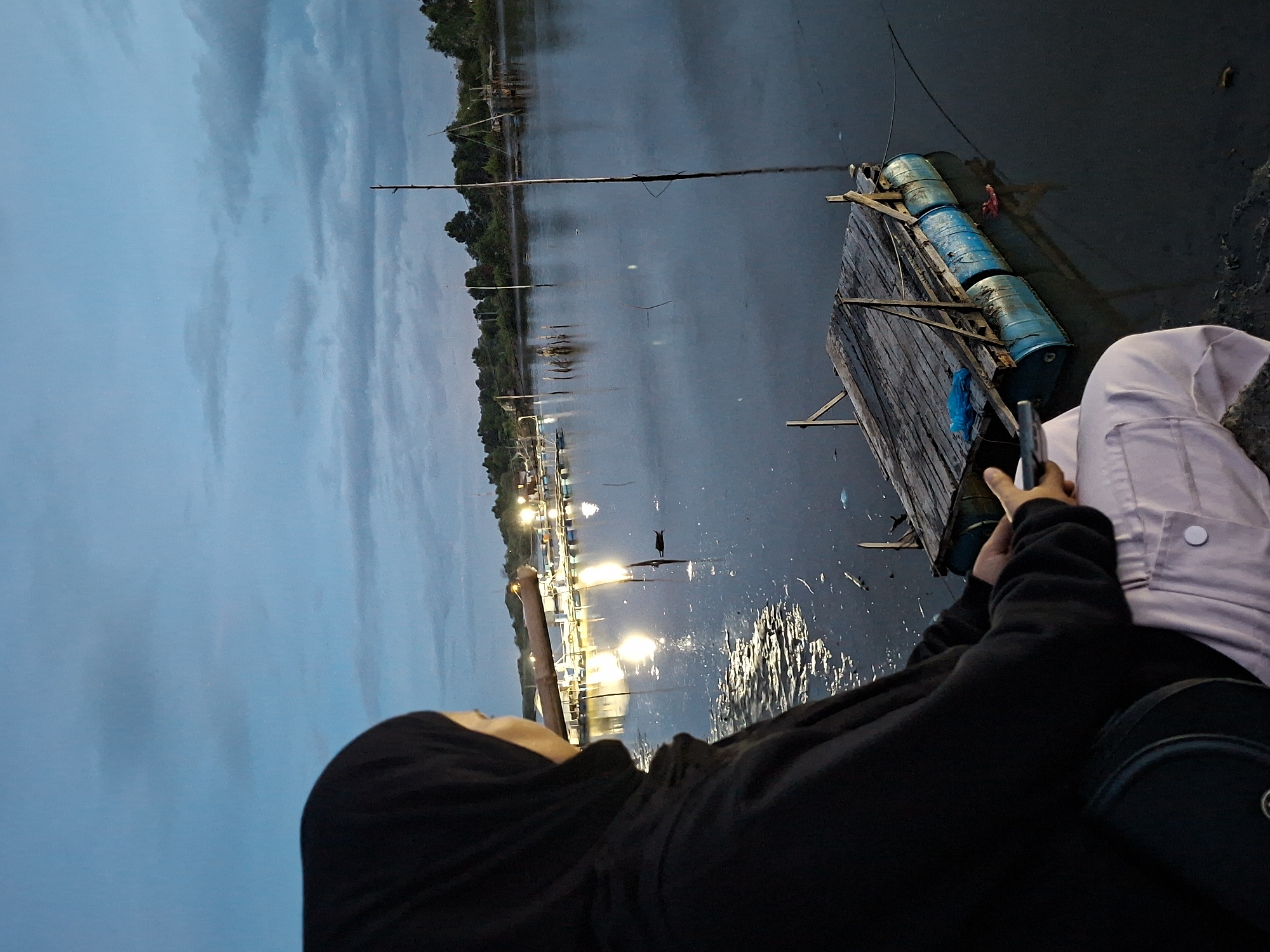 A person wearing a black hijab and a black hooded sweatshirt sits on a ledge, holding a smartphone in their hand, overlooking a body of water in Lhokseumawe, Indonesia. To the right is a platform made of wooden planks and supported by blue barrels. A long, thin fishing pole extends from the platform over the water. In the distance, there is a line of lights reflecting on the water. The sky above is overcast with blue-gray clouds.