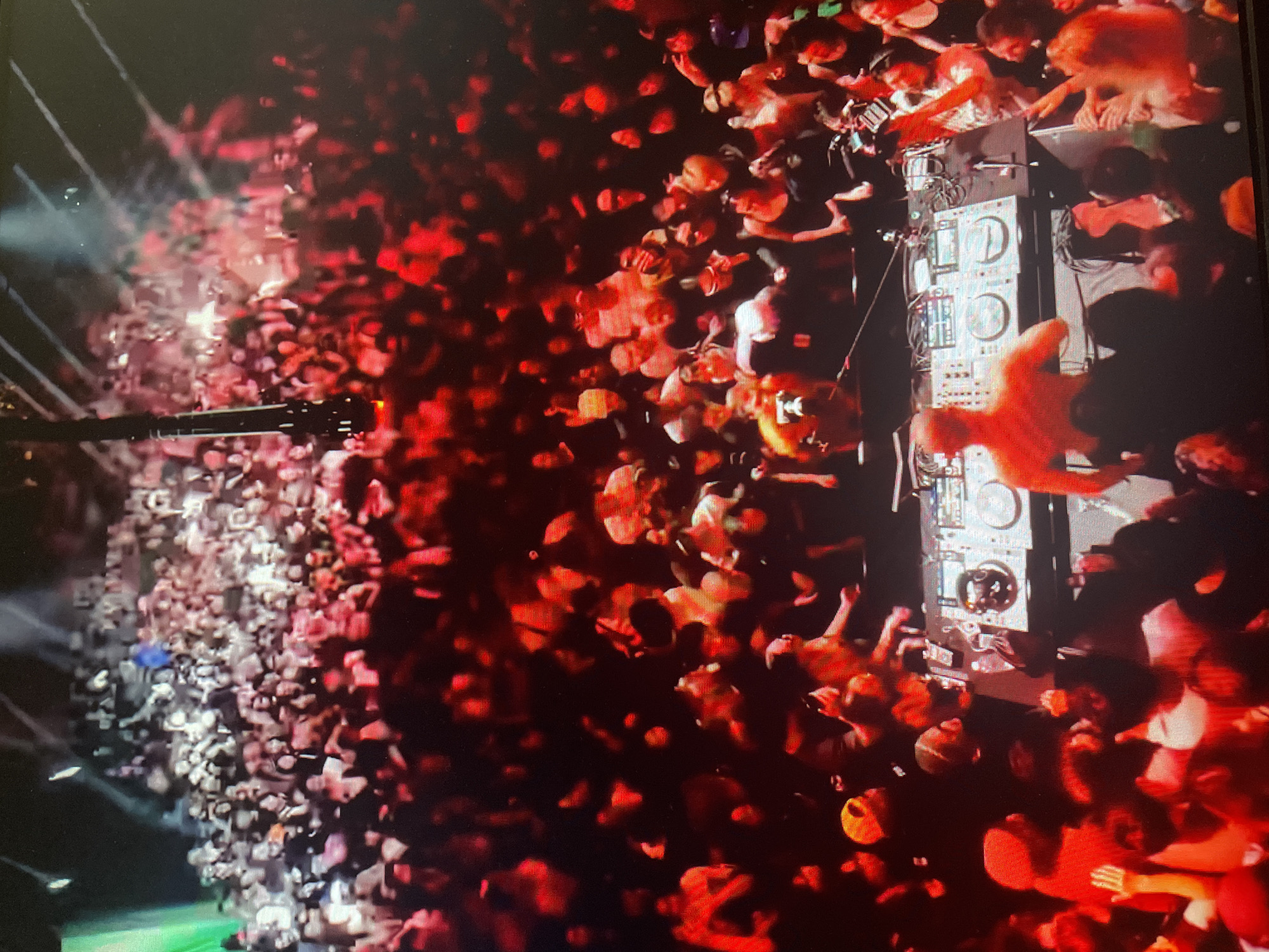 The image shows a high-angle, wide shot of a crowded nightclub or concert venue with a DJ setup in the foreground. A male DJ, with a muscular build and orange skin tone from the lighting, is positioned behind a DJ console, his back to the camera. The DJ console features multiple turntables, mixers, and screens. The crowd is densely packed, with people of varying ages and genders visible. Many are illuminated by red and white stage lighting, creating a vibrant and energetic atmosphere. Some individuals in the crowd are raising their hands, suggesting they are dancing or engaged with the performance. The overall environment appears to be an indoor venue with a dark ceiling and what appear to be stage lights or spotlights directed downwards. There are no clear indications of the city or country, including Unknown, Russia.