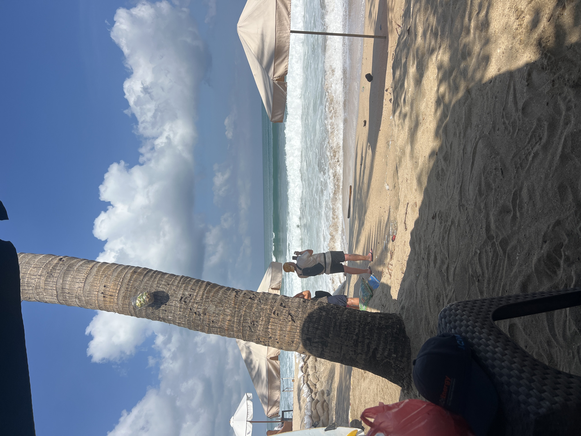 The image depicts a sunny day at Kuta Beach, Indonesia, with a focus on the sandy shore and the ocean. A large palm tree trunk dominates the left side of the frame, casting a shadow across the sand. In the foreground, a black cap with an orange logo rests on a dark plastic chair, with a red bag partially visible to its left.
Further into the scene, two individuals are standing on the beach. One, wearing a tan cap and shorts, is holding a phone up to take a photo or video of the ocean. The other person, partially obscured by the palm tree, is also facing towards the water.
The beach is lined with sandbags near what appears to be a low wall. Two large beach umbrellas, one white and the other beige, are set up further down the shore. The ocean is active with moderate waves crashing onto the beach, and the sky is a clear blue with scattered white clouds. Debris, including plastic bottles and other litter, is visible scattered on the sand. The overall impression is of a busy beach scene on a bright day.
