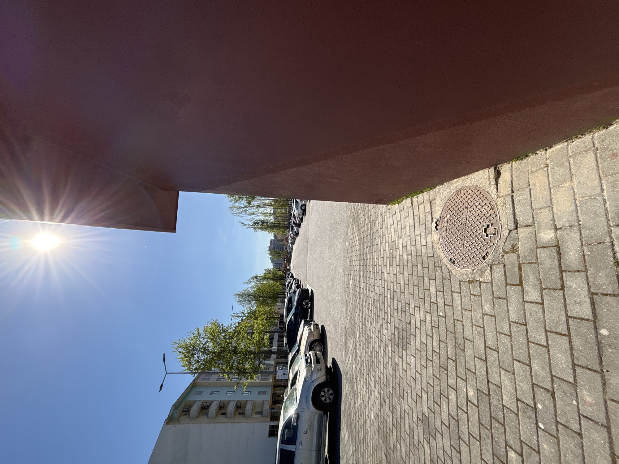 An exterior shot, slightly rotated, showcases a parking area adjacent to a building. Several cars are parked in a line, aligned with the curb. Above the cars and building, trees and a clear, bright sky are visible. A street light is attached to the building. A wide brick walkway is present, and a manhole cover is situated on the sidewalk. The building's structure has a horizontal dark-red panel above the parking area, creating a partial shadow. The location is suggested to be Mirandela, Portugal.