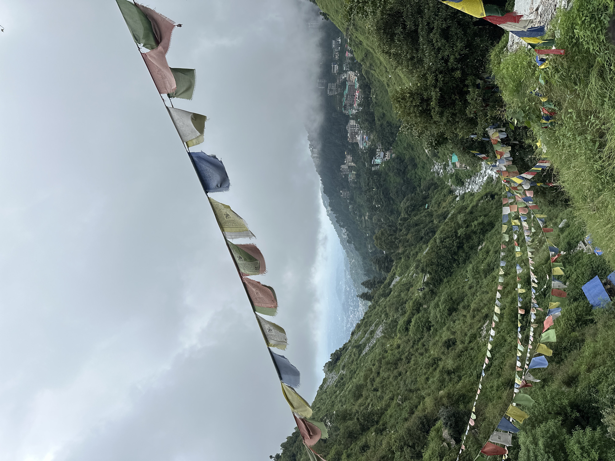 The image captures a scenic view of a mountainous landscape with prayer flags strung across the foreground. In the middle ground, a town is nestled within the valley, with buildings constructed on the slopes of the hills. The town appears to be Bhagsu, India, a popular hill station known for its spiritual sites and natural beauty. The prayer flags, typically found in Buddhist regions, suggest a spiritual or cultural significance to the location. The overcast sky and lush green vegetation indicate a possibly damp or humid climate. The overall scene evokes a sense of peace and tranquility, typical of mountain landscapes.