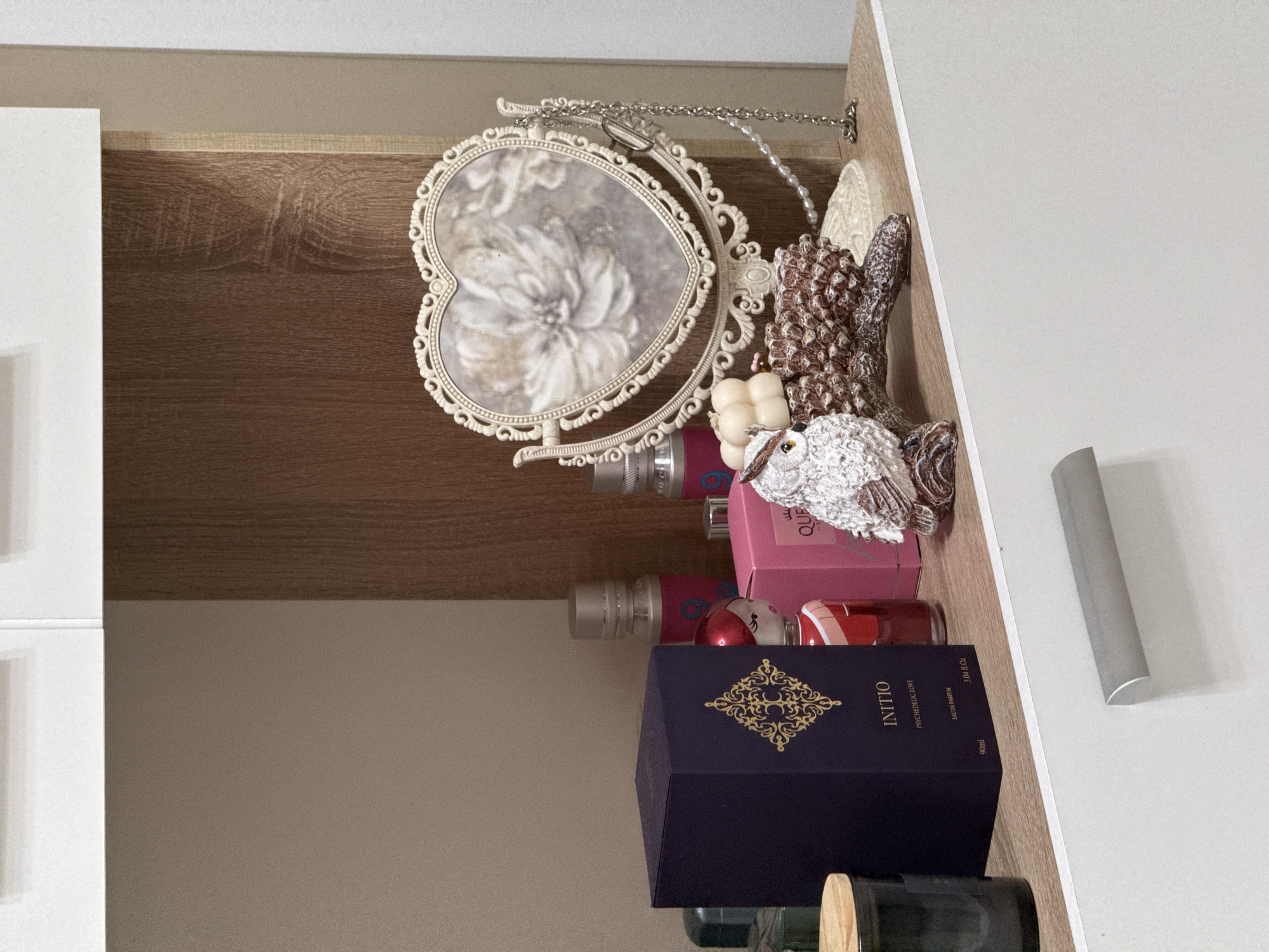 The image depicts a shelf containing a heart-shaped mirror with an ornate white frame, assorted perfume bottles and boxes, and decorative figurines. The perfumes include a dark blue box labeled "INITIO PSYCHEDELIC LOVE EAU DE PARFUM 90ml" and a pink box labeled "QUE". Additional items include a white owl figurine, a decorative pine cone, and a small white pumpkin. The shelf is likely located inside a cabinet, indicated by the white door panel on the left and the wooden panel behind the mirror. The precise location within the city of Unknown, Unknown is indeterminate from the image.