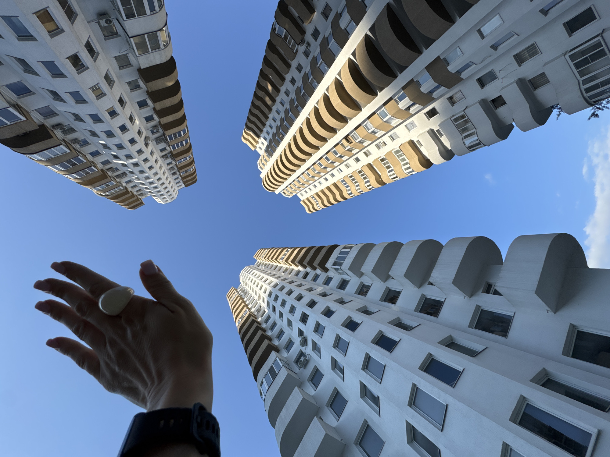 A hand wearing a large, light-colored ring and a dark smartwatch is held up against a clear blue sky, partially obscuring the view of three tall apartment buildings. The buildings are modern, with white facades and balconies or curved architectural elements in a light brown color. They are seen from a low angle, emphasizing their height. The sky is a uniform, bright blue, suggesting a sunny day. The image appears to be taken in Sochi, Russia, based on the architectural style of the buildings and common urban scenes in that region.