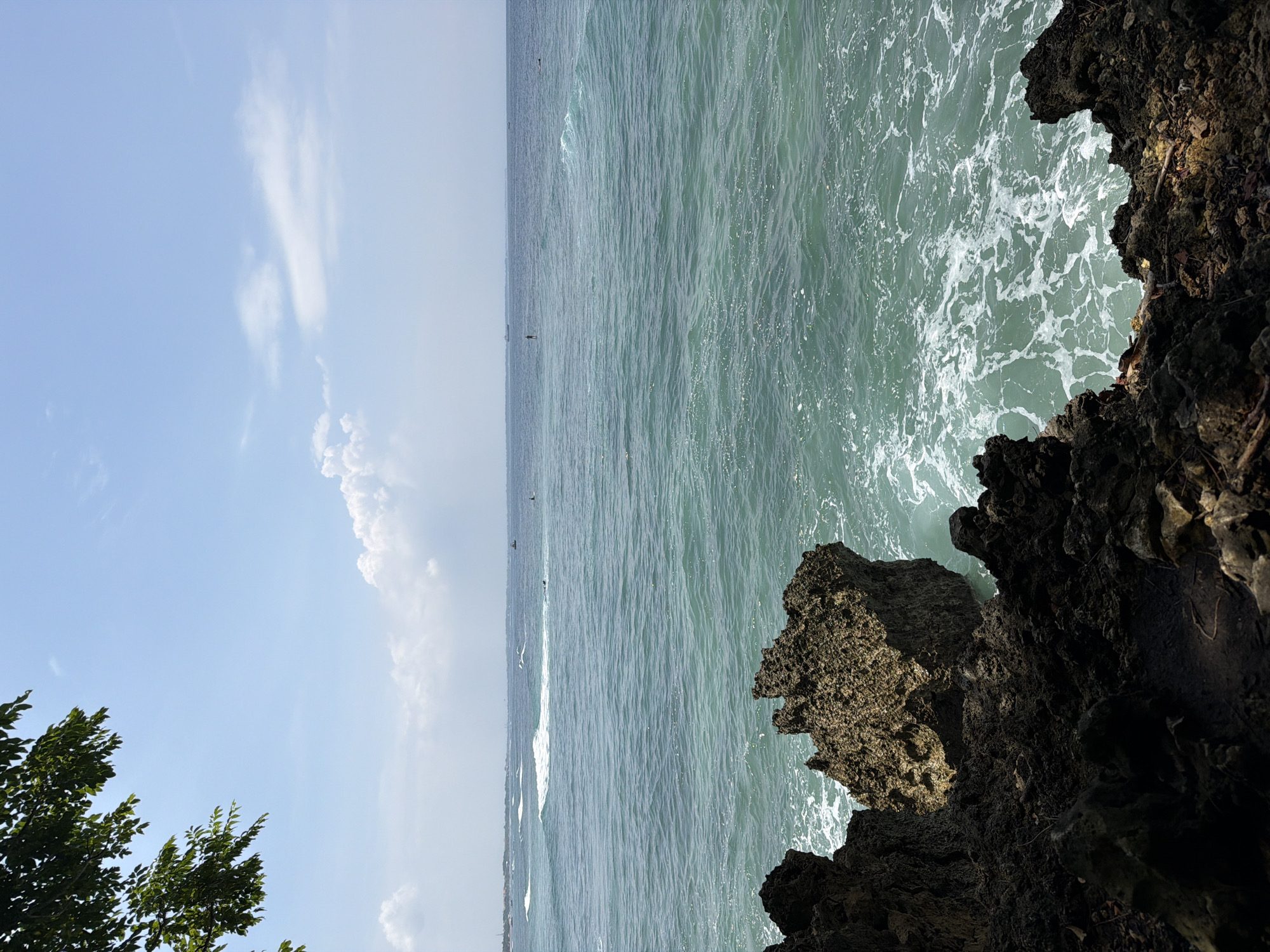 The scene depicts a coastal view in Nusa Dua, Indonesia, captured from an elevated position overlooking the ocean. The sky is clear blue with scattered white cumulus clouds. Dark green foliage from an unseen tree partially obscures the upper left corner of the frame. The immediate foreground is dominated by rugged, dark brown to black volcanic rock formations, with white ocean foam visible against their base. The ocean displays a gradient of turquoise to deeper blue-green, with multiple waves breaking into whitecaps across the surface. In the middle distance, at least two individuals are visible upright on the water, appearing to be on surfboards or paddleboards. Other dark, indistinct shapes in the water may represent additional people. A small, dark boat is present on the water to the far right, and a larger vessel is faintly discernible on the distant horizon. A hazy landmass is visible along the far left portion of the horizon.