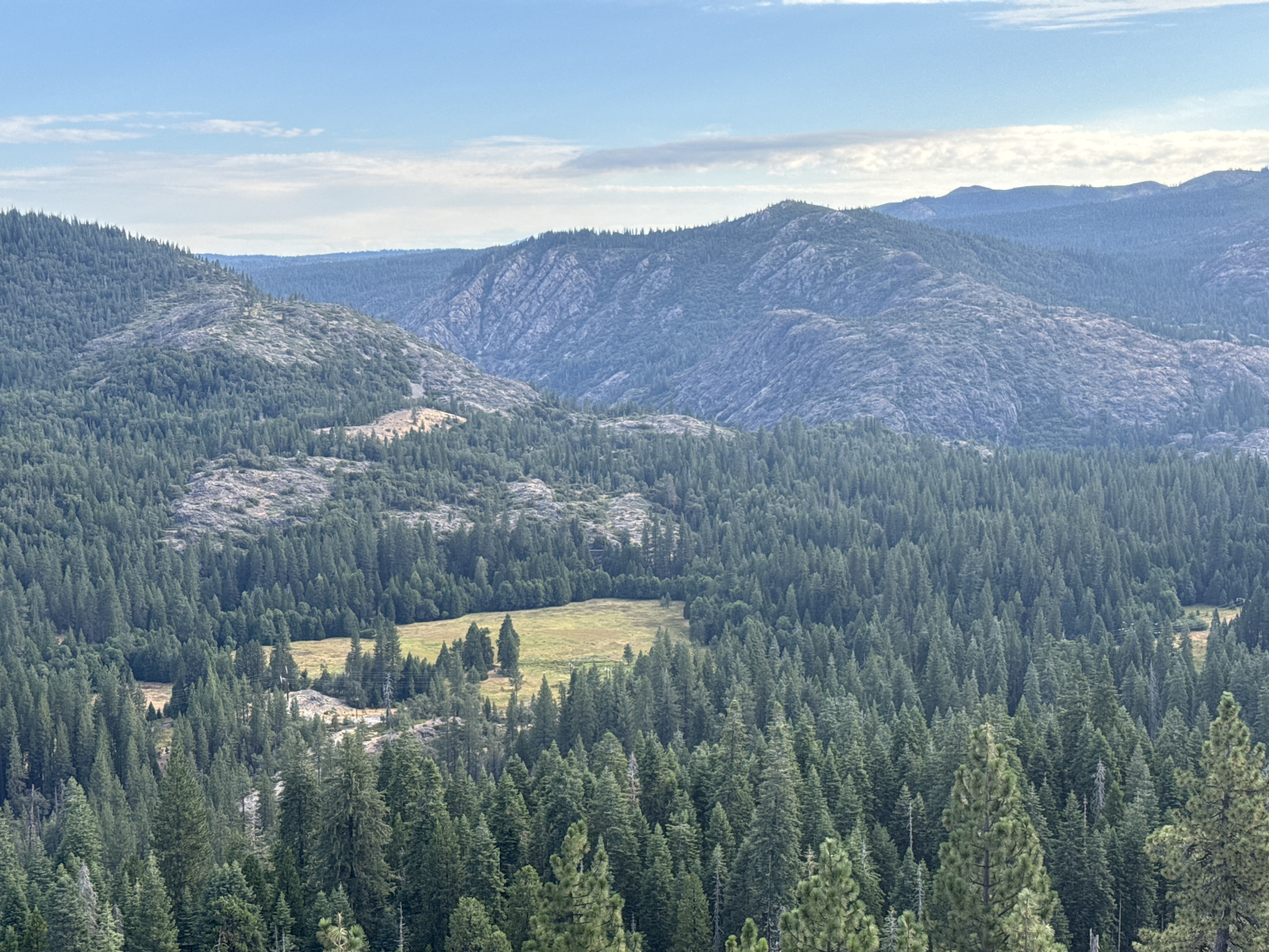 This is a wide-angle, eye-level shot of a forested mountain landscape in Emigrant Gap, United States. The scene is dominated by rolling hills covered in dense evergreen trees, primarily firs, with some rocky outcrops visible. In the mid-ground, a small, sunlit meadow breaks the uniformity of the forest, dotted with a few scattered trees. Further back, successive layers of mountains recede into the distance, becoming progressively bluer and more indistinct due to atmospheric haze. The sky above is a pale blue, with scattered, wispy white clouds.
The lighting suggests it is late afternoon or early evening, with the sun casting a warm glow on the meadow and creating subtle shadows within the forest. The weather appears clear and pleasant. There are no people or obvious man-made objects in the frame, emphasizing the natural, wild character of the setting. The overall impression is one of peacefulness and vastness, typical of a mountain wilderness. No text is visible in the image.