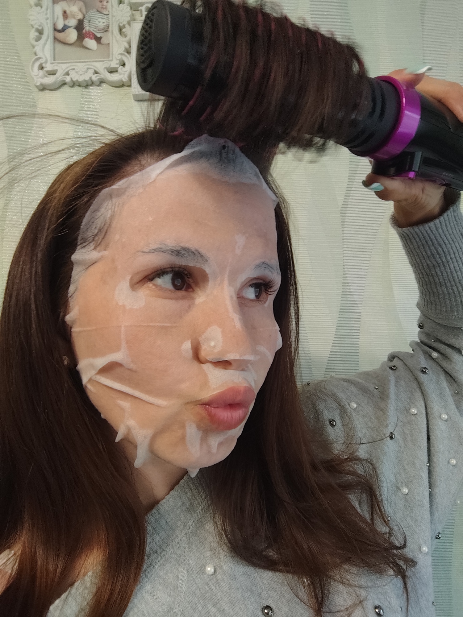 A woman is wearing a facial mask and using a hair dryer. The woman has brown hair and is wearing a gray sweater with pearl embellishments. She is holding a black hair dryer with a purple ring around the nozzle and has aqua-colored nail polish. The facial mask is white and covers most of her face. The background is a light green wall with a white framed picture of two babies. The wallpaper has a geometric pattern. It is not possible to identify the specific location of Фоки, Russia from the image.