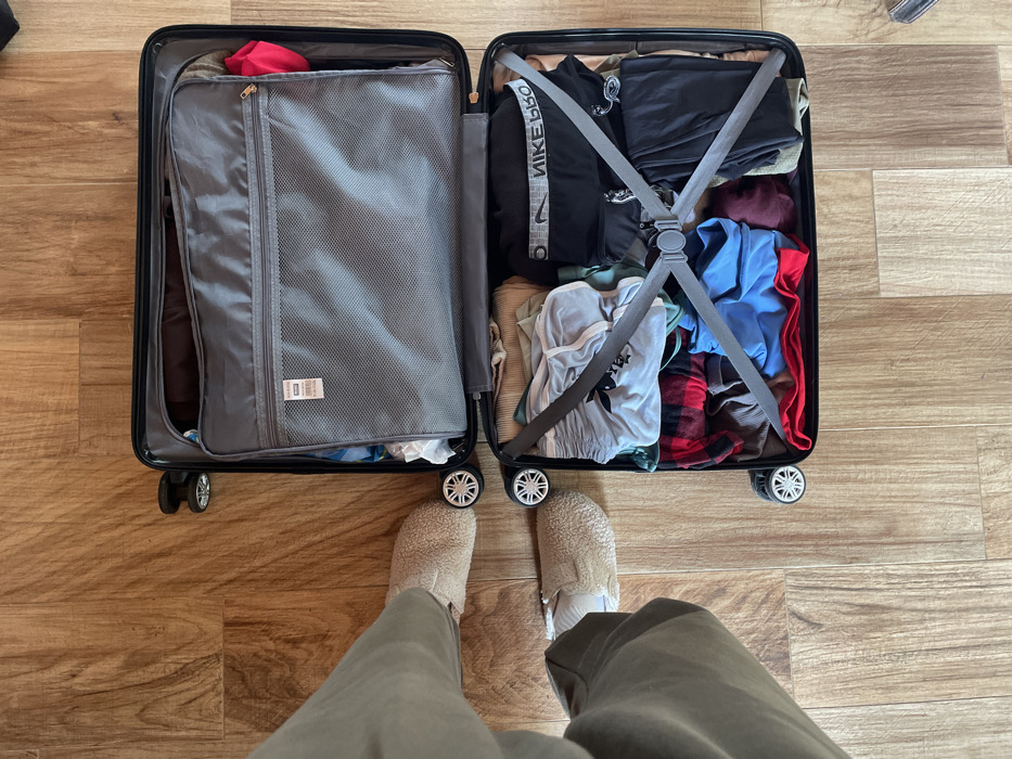 An overhead view displays the lower legs and feet of a person standing on a light brown wood-grain floor. The individual wears olive-green trousers and beige, fluffy slip-on shoes. Immediately in front of them lies an open, black hard-shell suitcase with spinner wheels. The suitcase is split into two sections: the upper section contains a grey fabric divider with a mesh zippered pocket, featuring a white tag marked "RFID BLOCKING," under which some clothes are visible. The lower section is filled with assorted packed clothing items in various colors, including red, blue, grey, black, and a red-and-black plaid pattern. A black athletic garment with "NIKE PRO" text is visible. These contents are secured by grey crisscross compression straps connected by a central buckle. The specific location within Monsummano Terme, Italy, cannot be determined from the visual information provided.