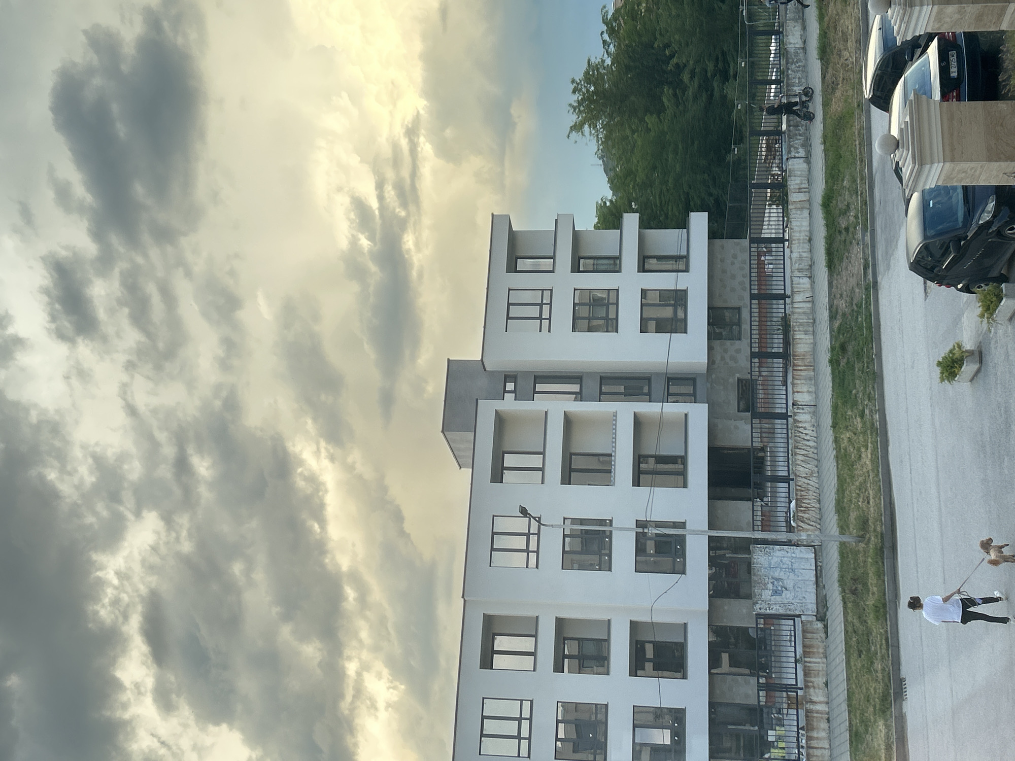 The image shows a modern, white apartment building in Plovdiv, Bulgaria. The building has a cubist design with recessed balconies and large windows. A sidewalk runs along the side of the building, bordered by a low metal fence and a strip of grass. Parked cars are visible along the sidewalk. A person is walking a dog on the sidewalk. The sky is overcast with dramatic cloud formations.