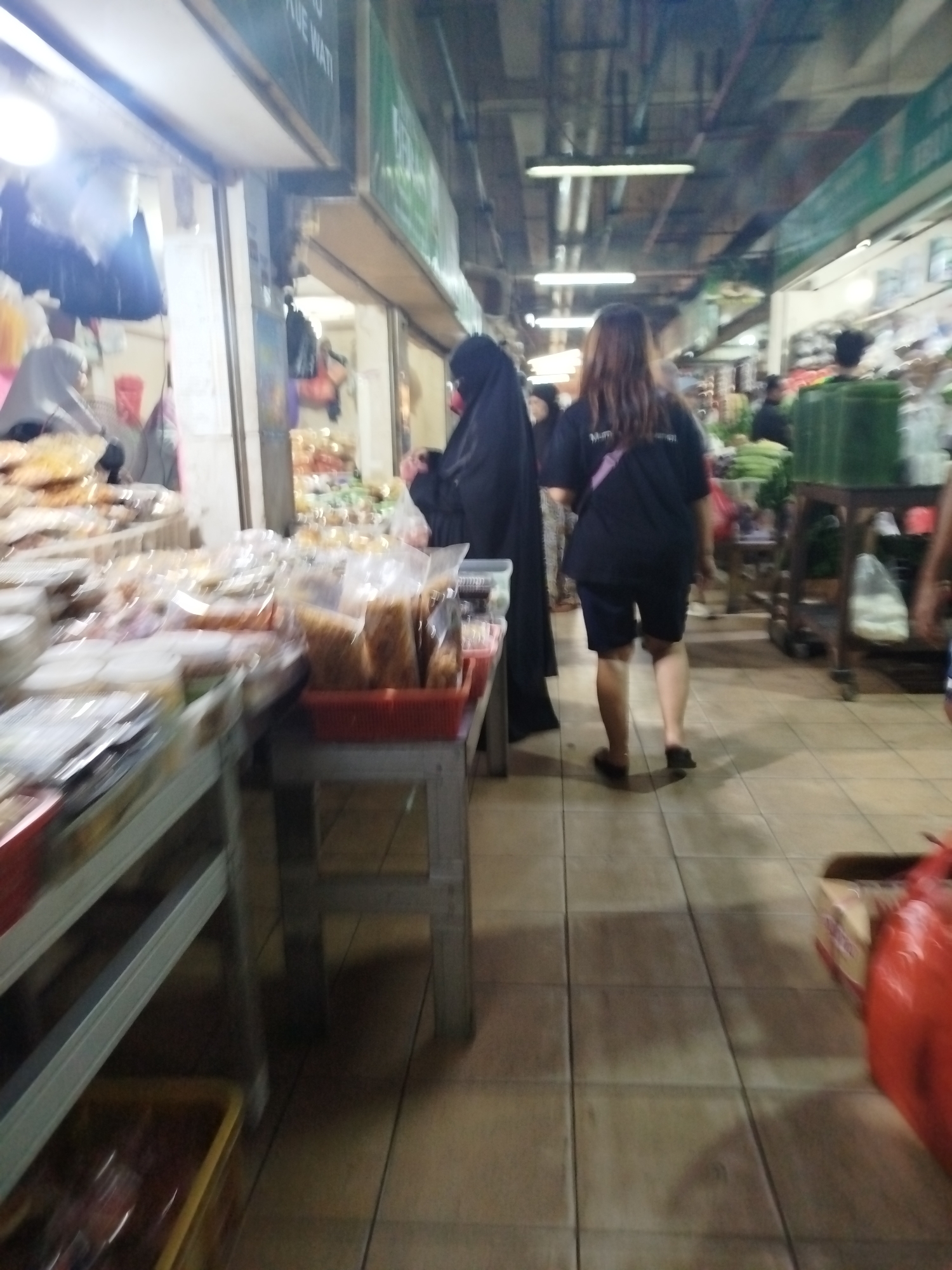 The image depicts an indoor market scene in Balikpapan, Indonesia, likely a traditional wet market or a food stall area within a larger market complex. The perspective is from a slightly low angle, looking down an aisle.
In the foreground, to the left, are several stalls with various food items, predominantly packaged snacks and possibly some baked goods, displayed in clear plastic bags and containers. These are arranged on metal shelves and tables. The overall impression is that of a busy retail environment.
Further down the aisle, two individuals are visible. On the left, a person wearing a black hijab and abaya is standing near a stall. To their right and slightly ahead, a woman with long brown hair is walking away from the camera. She is wearing a black t-shirt with a logo on the back and dark shorts, with a pink strap bag slung across her chest. Her attire suggests a casual shopping trip.
The environment is characterized by tiled flooring, overhead lighting, and the general clutter and varied merchandise typical of a bustling market. The lighting appears to be artificial, with some light sources visible in the ceiling. The overall image has a somewhat blurred quality, suggesting motion or a less than optimal camera. The stalls visible behind the main subjects show more displays of food items, some in baskets and plastic containers, with a green display on a wooden cart to the far right, possibly containing vegetables or other produce. The presence of large red plastic bags on the far right suggests merchandise or packaging being transported.