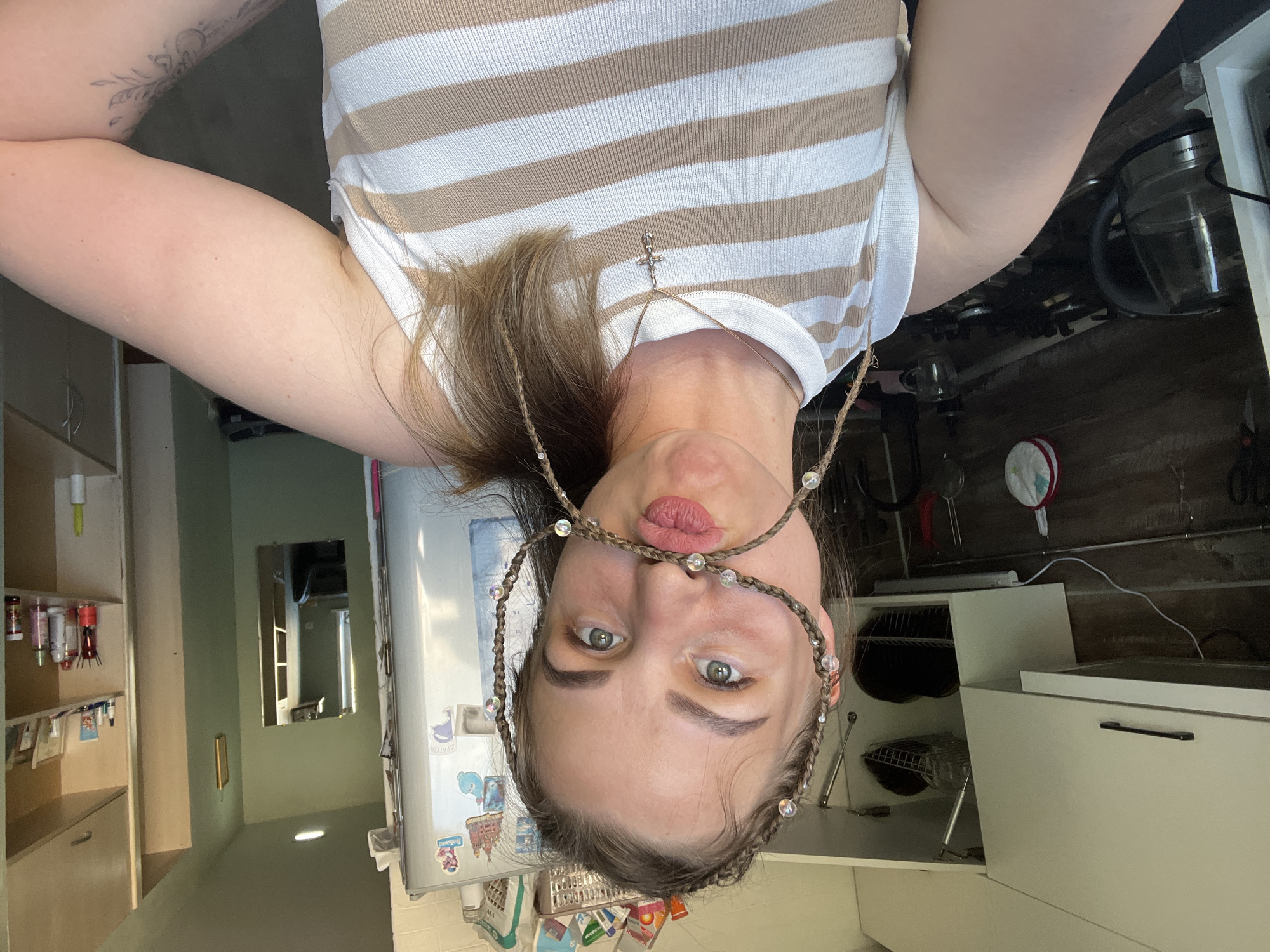 A woman is oriented upside down in the image frame. She is wearing a horizontally striped white and beige shirt. A braided headband with interspersed clear beads frames her face, connected by a thin necklace with a cross pendant. Her eyes are open and looking at the camera. The background includes a kitchen setting with white cabinets, a refrigerator covered in magnets and stickers, and cooking utensils hanging on the wall. A mirror reflecting another room can be seen in the distance. There is no information to indicate the location is Izhevsk, Russia.