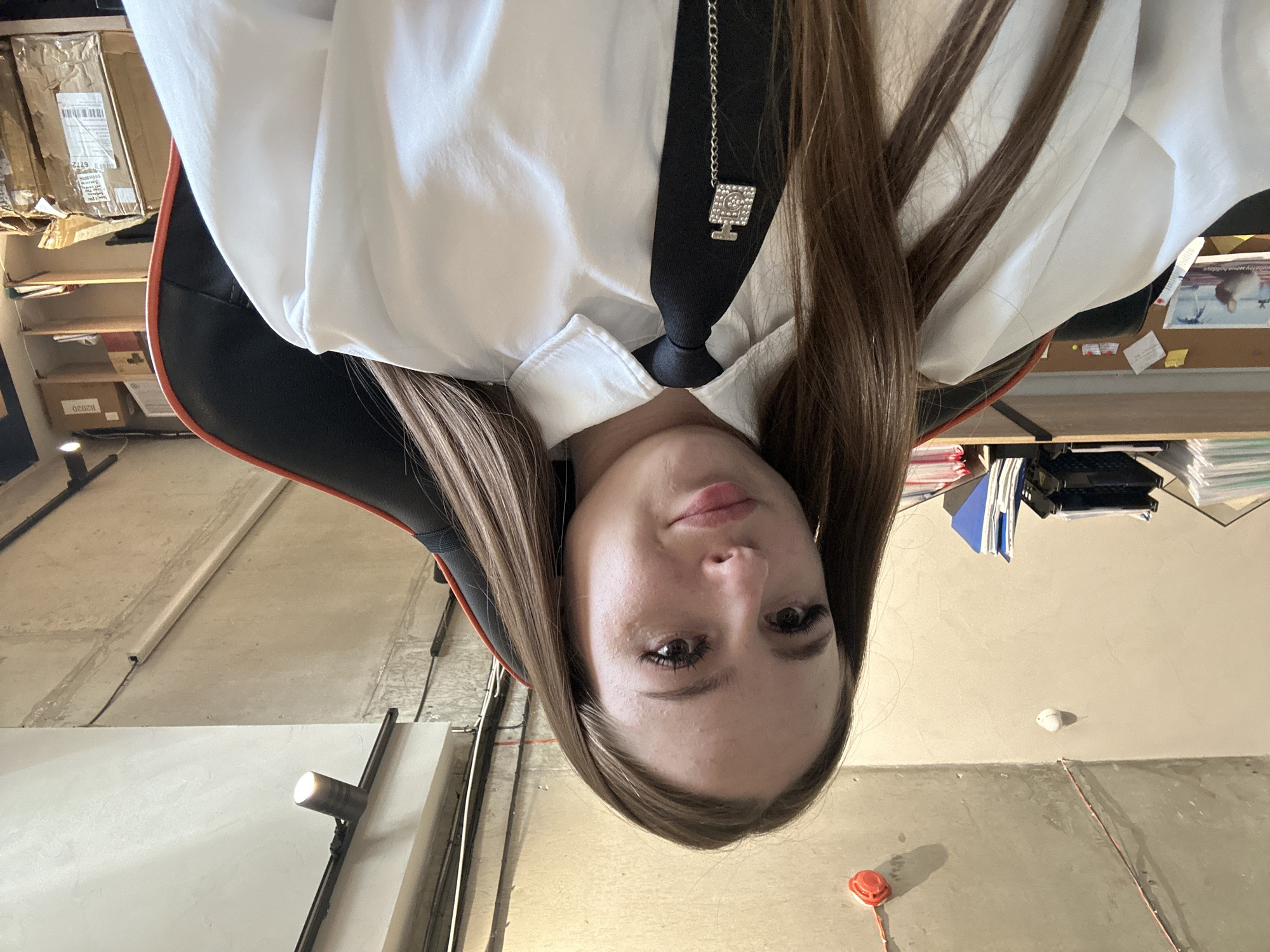 A young woman with long, brown hair is seated in a gaming chair, looking at the camera. She is wearing a white collared shirt and a black tie with a decorative silver pendant shaped like a perfume bottle. The background shows shelves filled with boxes and papers, and a concrete ceiling with exposed wires and light fixtures. The scene appears to be an indoor office or workshop.