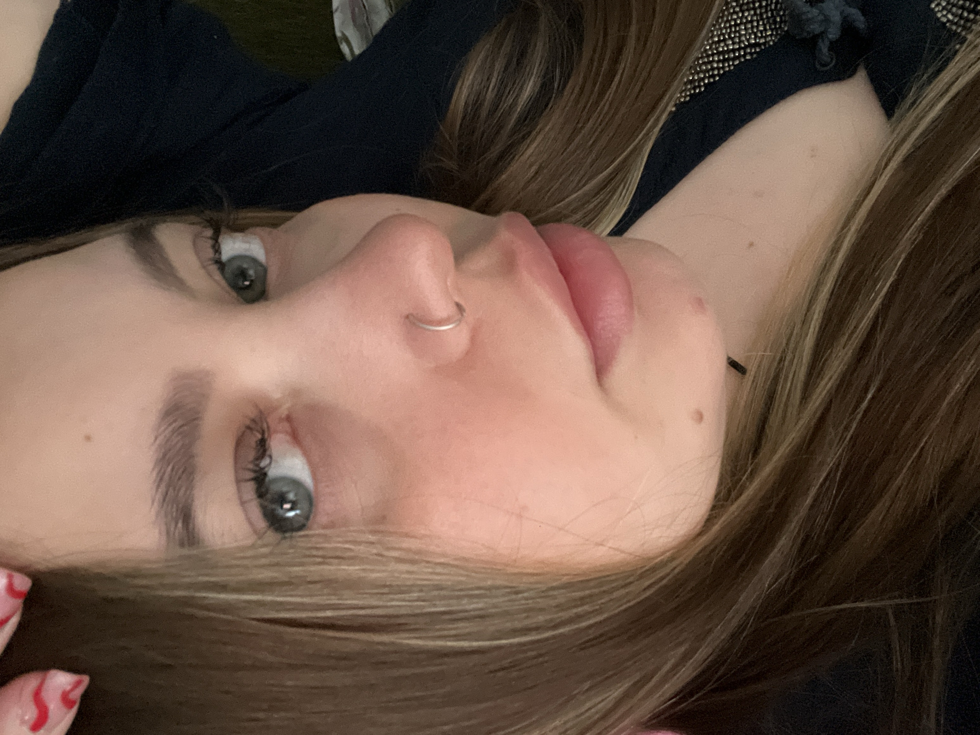 A close-up, slightly off-kilter shot of a young woman's face. She has light brown hair, fair skin, and blue-grey eyes. She wears a small silver nose ring. The background is blurred and includes dark clothing or fabric.
The image contains no information related to the location in the city of Novopokrovskaya, Russia.