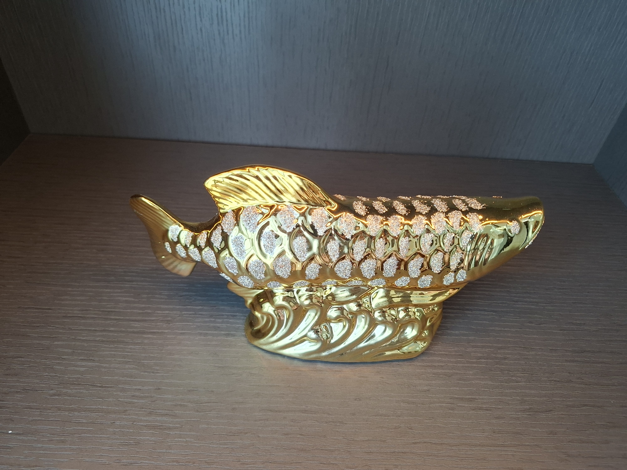 An ornate, golden fish figurine with textured scales rests on a wooden surface. The figurine is positioned on a shelf with a dark, textured wall behind it. The figurine has a glossy finish with inlaid white accents on its scales, and it sits on a base designed to resemble stylized waves. The lighting appears to be soft, casting gentle reflections on the golden surface. There are no people or animals present in the image. The scene is static and appears to be an indoor display. The location in West Jakarta, Indonesia is not discernable from the image.