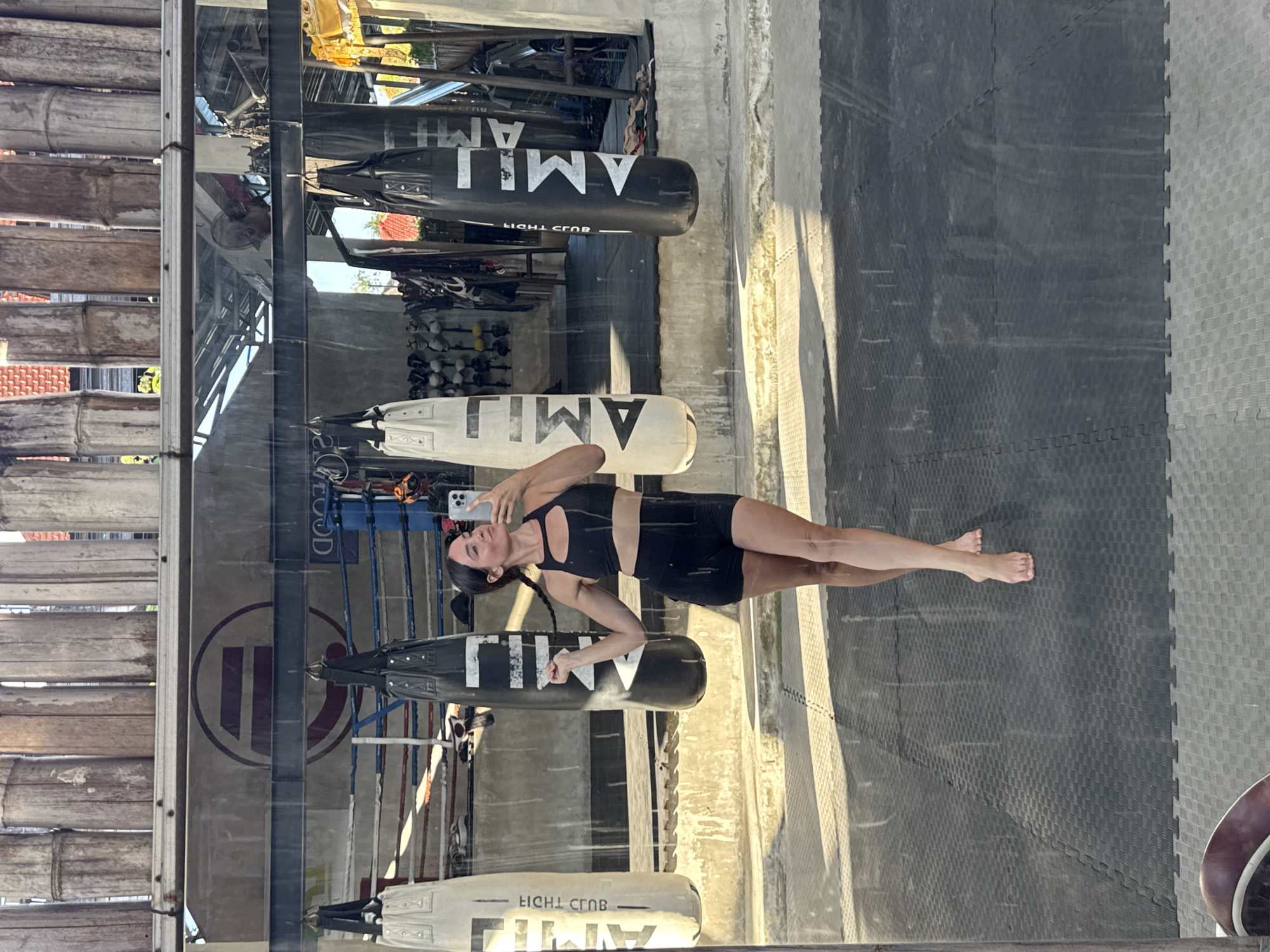 A woman in athletic wear is taking a mirror selfie at a gym. She is wearing a black sports bra and black shorts. She has her hair in braids and is holding a phone in her right hand. She is barefoot. In the background, there are several punching bags hanging, a boxing ring, and other gym equipment. The gym appears to be open-air, with a wooden ceiling structure and a concrete floor. The lighting suggests it is daytime. The location is identified as a gym in Kuta Selatan, Indonesia.