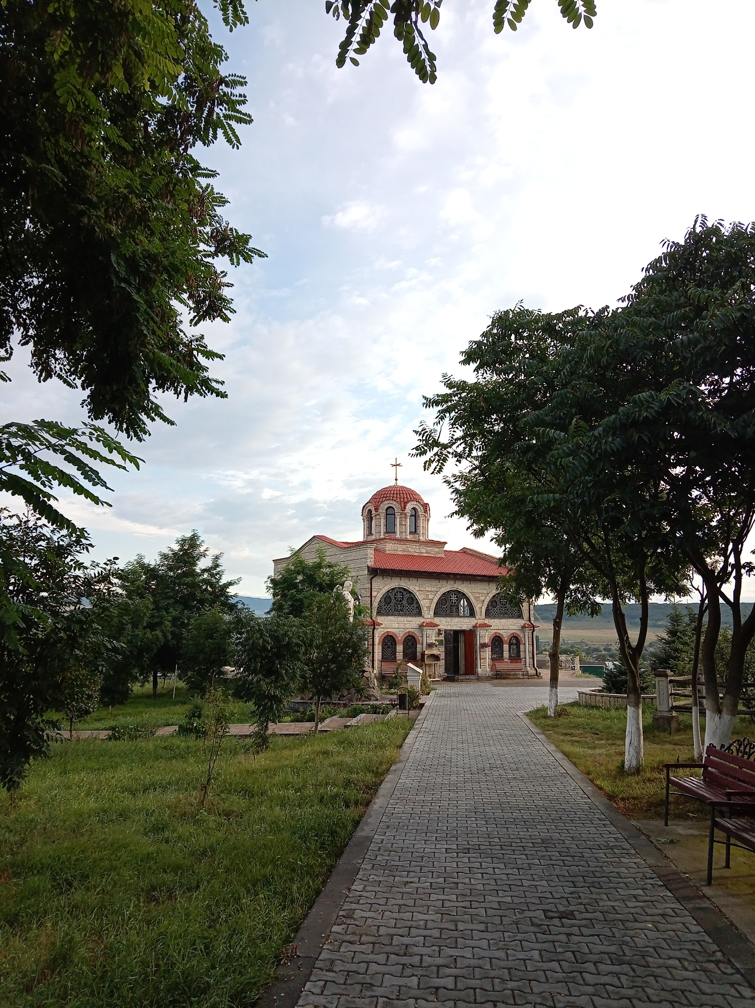 The image depicts the Church of the Great Martyr George in Yessentuki, Russia, on a slightly overcast day. The church, constructed of stone with a red-tiled roof and a prominent dome topped with a cross, is centrally located. A paved walkway leads to the church entrance, flanked by green grass and trees. To the right of the walkway, a wooden bench is visible. The background shows a gentle slope with sparse vegetation and distant hills. The sky is a mix of light clouds and blue. There are no people present in the image.