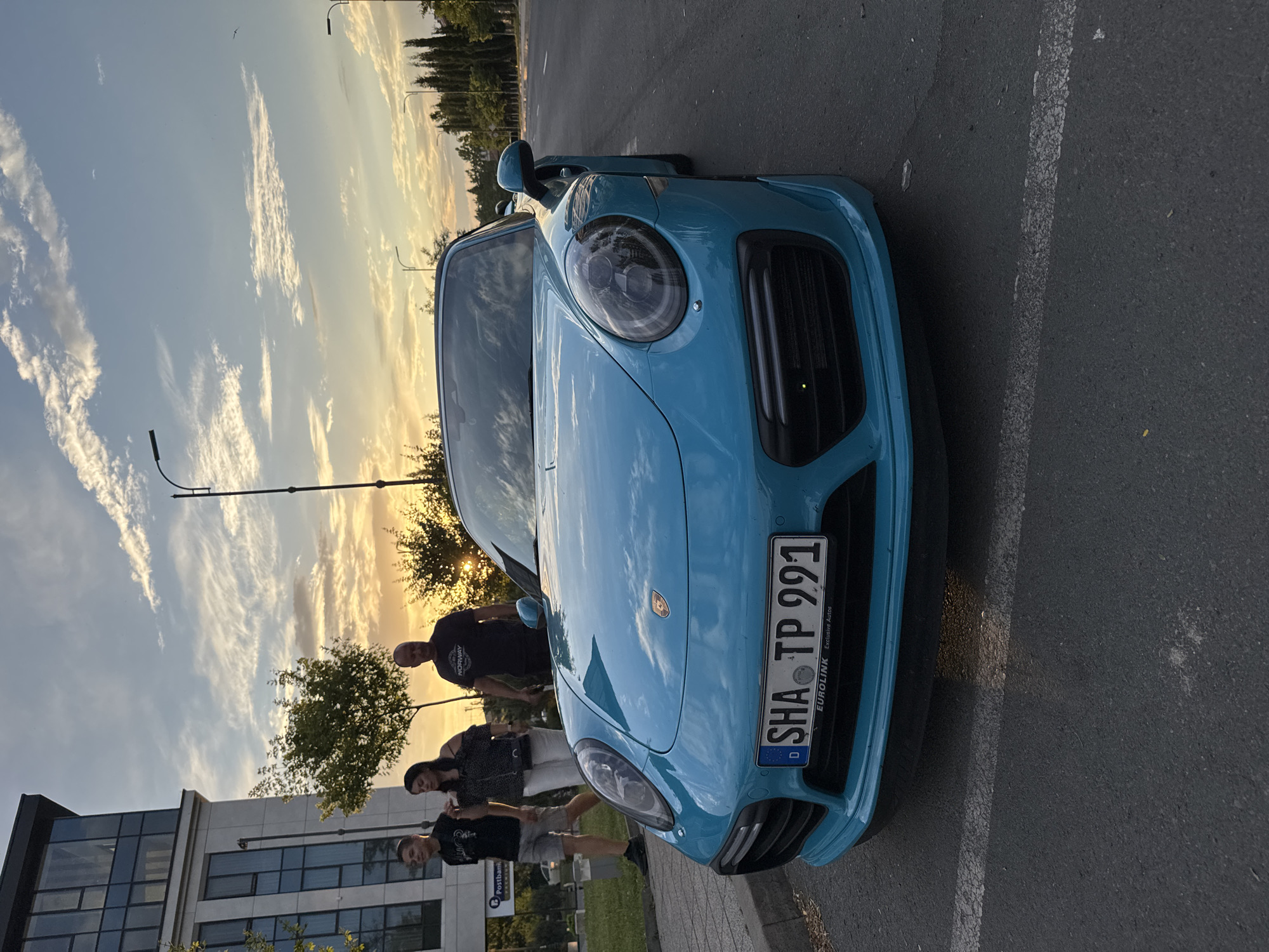 The media file captures a group of people standing near a bright blue Porsche on a street in Burgas, Bulgaria. The scene is set during sunset, with a dramatic sky filled with wispy clouds illuminated by the golden light. Trees and streetlights are visible in the background, suggesting an urban or suburban environment. The car's license plate is clearly visible and reads "SHA TP 991". Text below the plate indicates "EUROLINK Exclusive Autos". The people appear to be casually dressed, and the overall mood seems relaxed, possibly a social gathering or a moment to admire the car. The building in the background appears to be a modern office building, possibly a bank given the "Postbank" sign. The sun is low on the horizon, casting long shadows and bathing the scene in a warm, orange glow, characteristic of a late summer or early autumn evening.