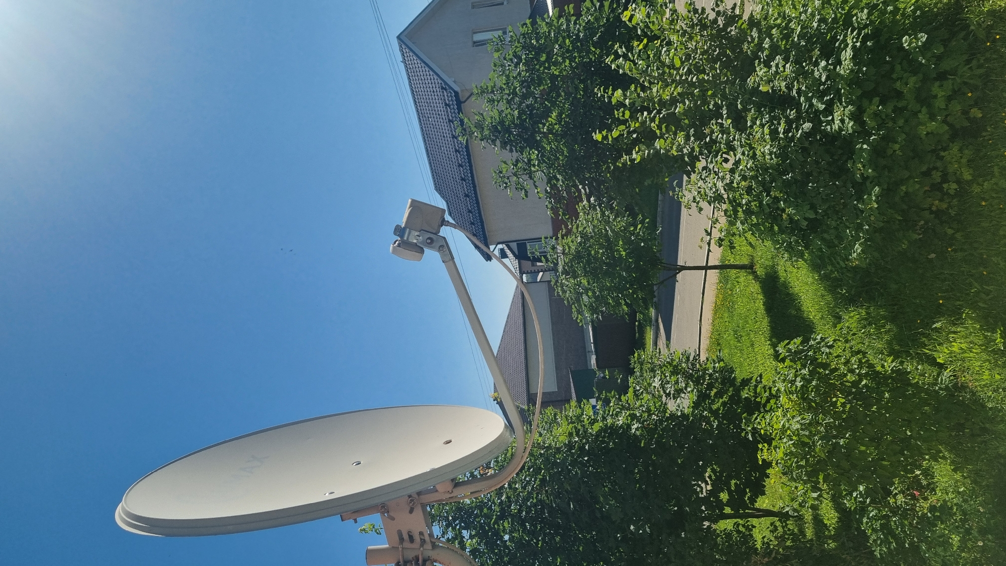 In the town of Verzilovo, Russia, on a clear, sunny day, a satellite dish is mounted on a structure, angled towards the bright blue sky. The dish, a light color, has the word "MAX" visible on its surface. It is supported by a metal arm extending from a base. Behind the dish, lush green trees and bushes create a natural foreground. In the background, residential houses with dark roofs are visible, along with a paved road and more green spaces. The overall scene depicts a typical suburban or rural environment on a pleasant day.