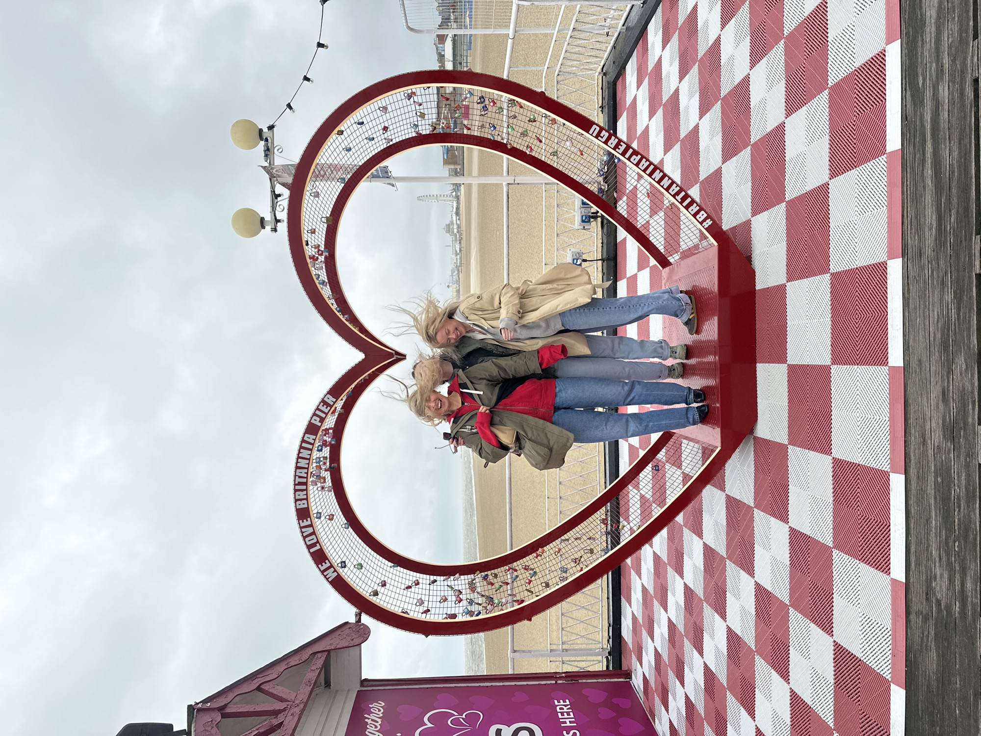 This image captures three women standing in front of a large, red, heart-shaped love lock structure on Britannia Pier in Great Yarmouth, United Kingdom. The structure, positioned on a red and white checkered floor, is inscribed with "WE LOVE BRITANNIA PIER" across its upper curve and "#BRITANNIAPIER" on its lower right side, and is adorned with numerous colorful padlocks.
The three women are positioned within the heart frame, their blonde hair visibly windblown to the right, suggesting breezy conditions. From left to right: the first woman wears a green jacket over a red top and blue jeans, holding a light-colored handbag. The central woman is partially obscured but appears to wear a dark top. The third woman wears a light beige trench coat over a grey hoodie and blue jeans. All three women are smiling or laughing, looking towards the viewer.
The pier's floor is composed of red and white interlocking square tiles with a textured pattern, bordered by dark wooden planks in the foreground. To the left, a section of a pink building with white trim is visible. In the background, a wide, sandy beach stretches towards the grey, choppy sea under an overcast sky. Distant coastal structures, including a Ferris wheel, are visible on the horizon. Overhead, two yellow globe-shaped light fixtures and a string of exposed light bulbs are present.