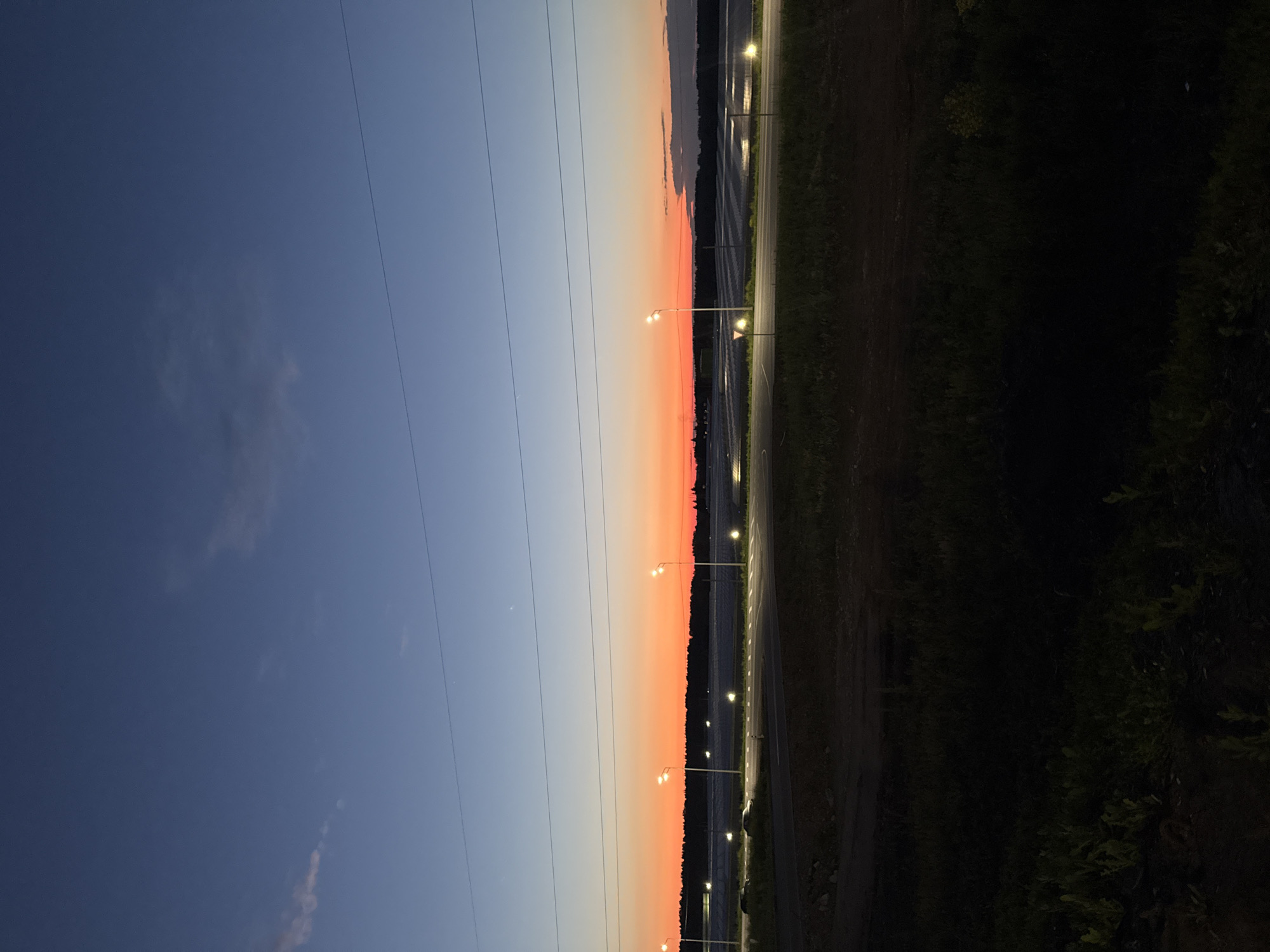 A vertical, low-angle, full outdoor shot captures a twilight landscape at dusk with a gradient sky transitioning from deep blue at the top to vibrant orange and red near the horizon. Three power lines stretch horizontally across the upper third of the image. Below the sky, a line of trees forms a dark silhouette against the colorful sky. In the middle ground, a road is visible with several streetlights illuminating the area. Two cars are parked or moving slowly on the road. The foreground is a dark, silhouetted field of grass and some scattered foliage. The overall impression is one of a rural or semi-rural environment at the edge of a city during twilight.